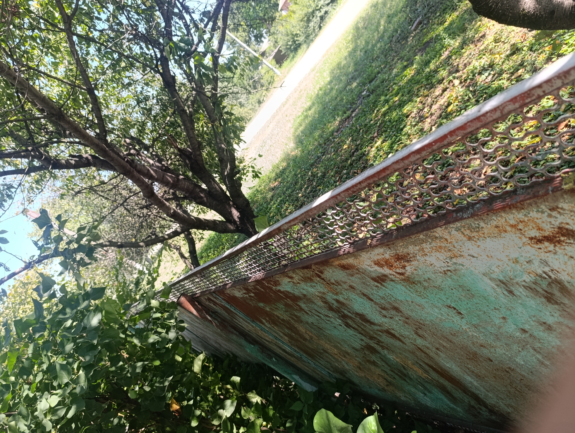 The image depicts an outdoor scene with lush green foliage from trees and bushes dominating the foreground and left side of the frame. A weathered, rusted metal structure, possibly part of a fence or barrier, runs diagonally across the lower right portion of the image, displaying streaks of teal and brown paint. Behind the metal structure, a sun-drenched grassy area slopes upwards, with a faint path or road visible in the distance, bathed in bright sunlight and casting dappled shadows. The background reveals more trees and greenery, suggesting a park or a natural setting. There are no people or animals visible in the image. The setting is likely residential or recreational, with a sense of overgrown nature.