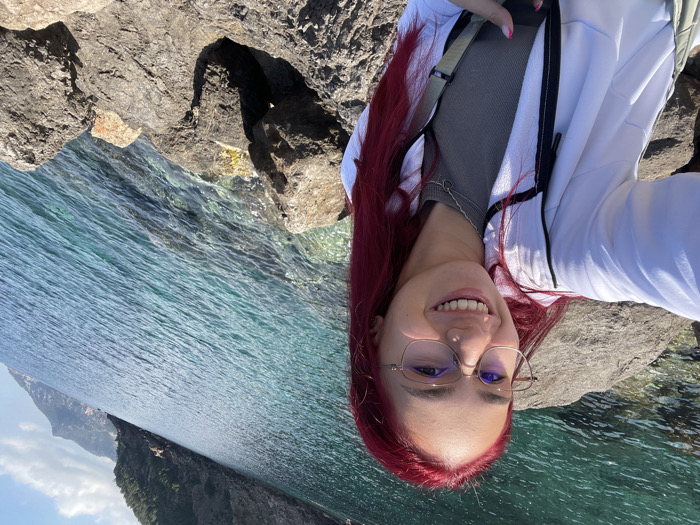 A young adult female is depicted in a selfie-style photograph taken on a sunny day in a coastal area near Valldemossa, Spain. She has long, bright red hair, wears eyeglasses with light-colored frames, and has a nose piercing on her left nostril. She is smiling broadly, showing her teeth. Her attire consists of a white zippered jacket with black accents over a grey or olive-green ribbed top, and a thin silver-colored necklace. Her fingernails are painted pink. The woman is positioned on a rocky shoreline, with dark, irregular rocks forming the immediate foreground. The background features clear, tranquil turquoise-blue sea water exhibiting ripples and visible submerged elements. Beyond the water, steep, vegetated cliffs rise prominently, consistent with the Serra de Tramuntana coastline, under a partly cloudy blue sky.