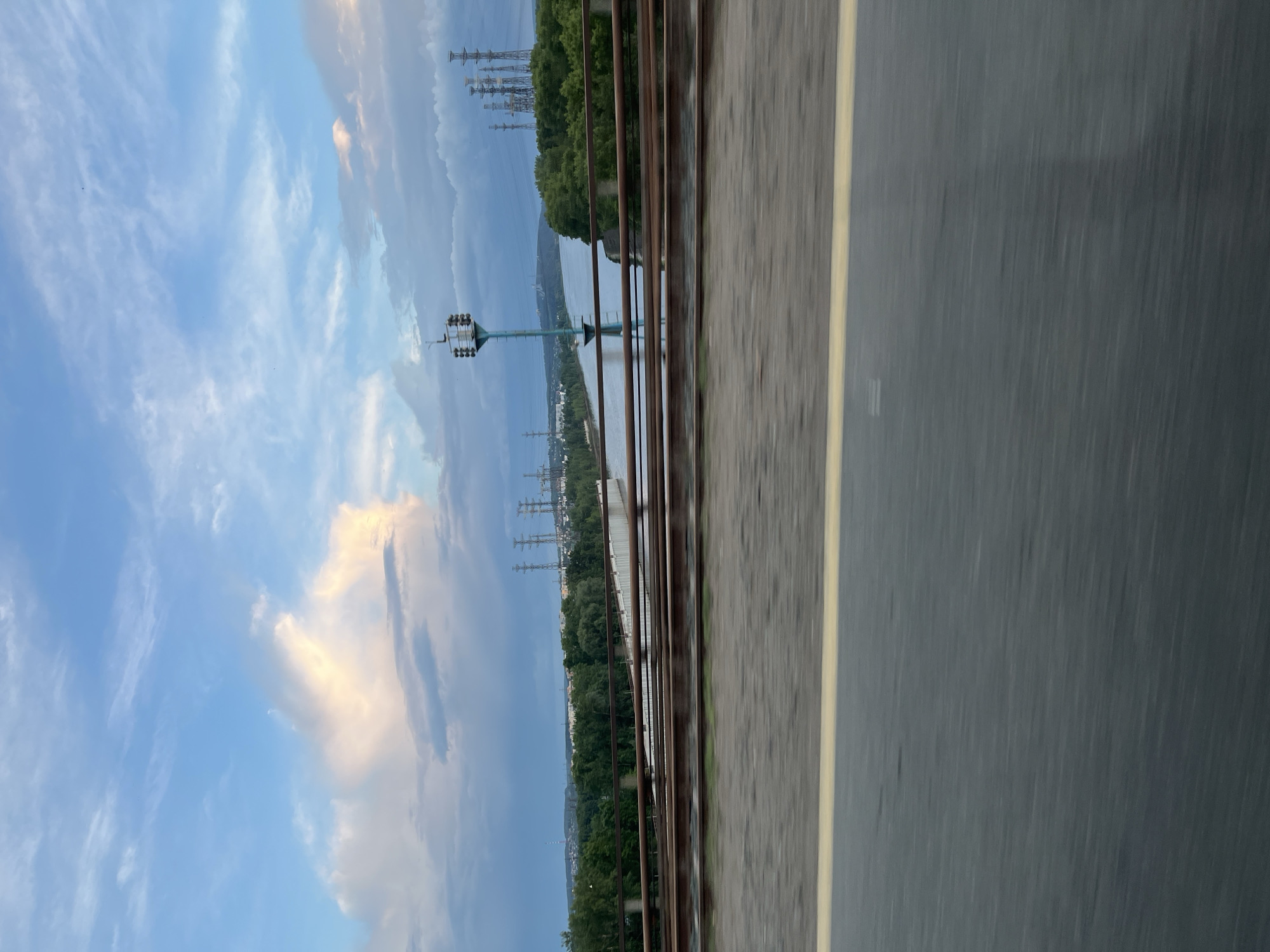 The image shows a view from a bridge in the city of Chaikovsky, Russia. The bridge appears to be a vehicle bridge, with a gray asphalt road taking up the lower right portion of the frame. A yellow line marks the road's edge. A brown metal guardrail runs parallel to the road. Beyond the guardrail, the cityscape is visible. The Kama River flows between the bridge and the city. Electric transmission towers stand on both sides of the river. A light blue sky with white clouds fills the upper left portion of the frame. Green trees line the river banks and the edges of the city.