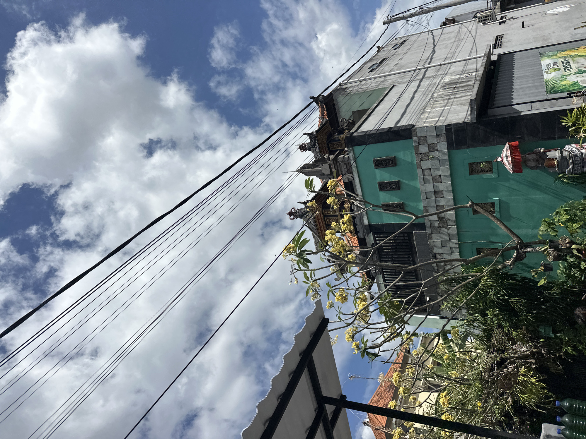 A photograph taken from a low angle on a sunny day shows a building with a turquoise and gray facade, topped with traditional Balinese roof decorations. The building is partially obscured by a frangipani tree with yellow flowers and lush green foliage. Overhead, multiple black electrical wires stretch diagonally across the frame against a blue sky dotted with white cumulus clouds. To the left, a white corrugated awning extends outwards. In the foreground, several plastic bottles are visible. The scene appears to be a street in North Kuta, Indonesia, given the architectural style and vegetation.