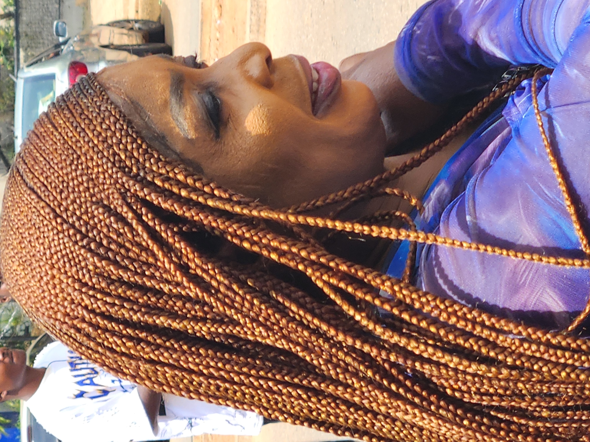 An image shows a close-up, side profile of a Black woman with micro braids in her hair. She is wearing a shimmering purple garment. In the background, another person is visible wearing a white t-shirt with the word "CAUTION" printed on it. A silver car is also parked in the background. The scene appears to be outdoors, possibly on a street, given the pavement and the presence of vehicles. The lighting suggests it is daytime.