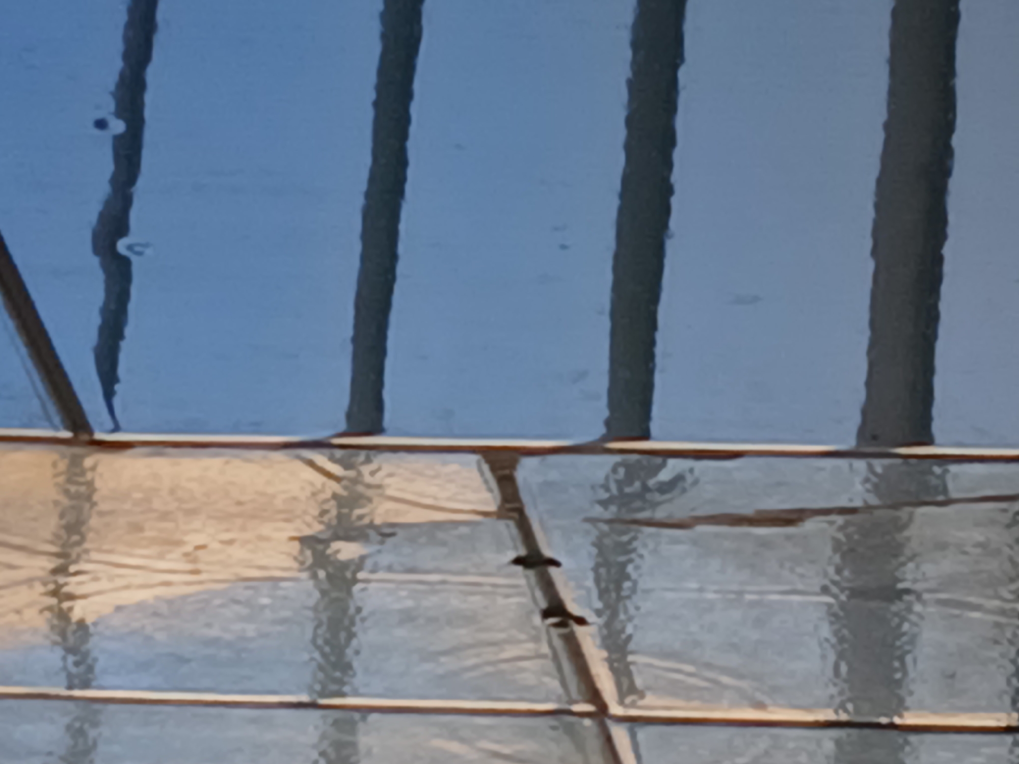 The image shows a section of a glass roof or ceiling, likely part of a building structure. The glass panels are separated by dark, vertical support beams. There are some water droplets present on the glass surface. The lower half of the image reveals a reflection of the sky above, which appears hazy. The location is not discernable from the image. There are no people visible in the image, so the location in the city of Ciudad Guayana, Venezuela is unable to be verified.