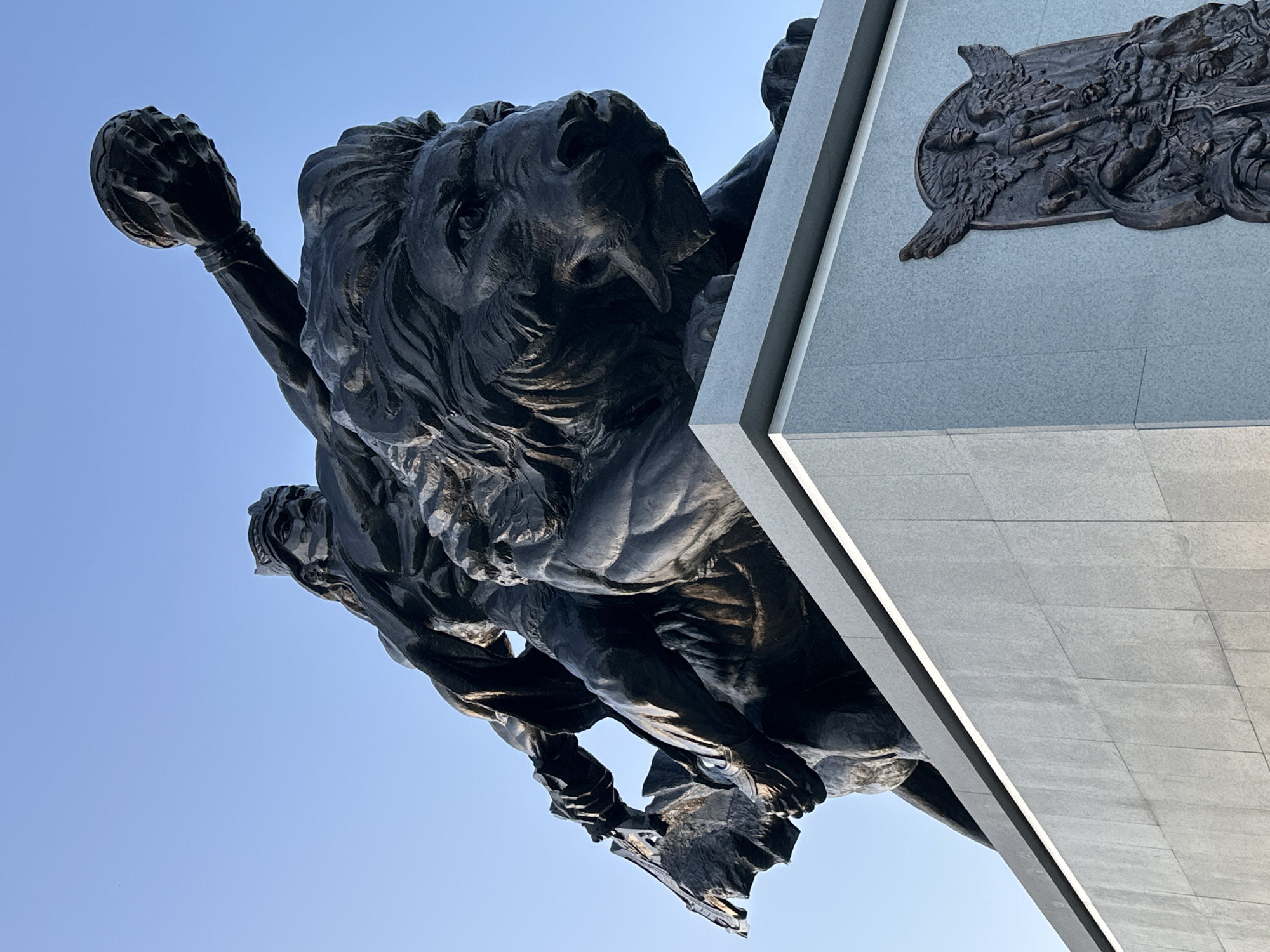 The image shows a bronze sculpture of a lion straddling two figures, seemingly humans, atop a stone monument. The lion holds an object in its raised paw, while the figures beneath appear to support the lion's weight. The monument has a light gray, tiled exterior with a bronze plaque depicting figures in relief. The sky is clear and blue. The location is near the National Pantheon in Lisbon, Portugal.