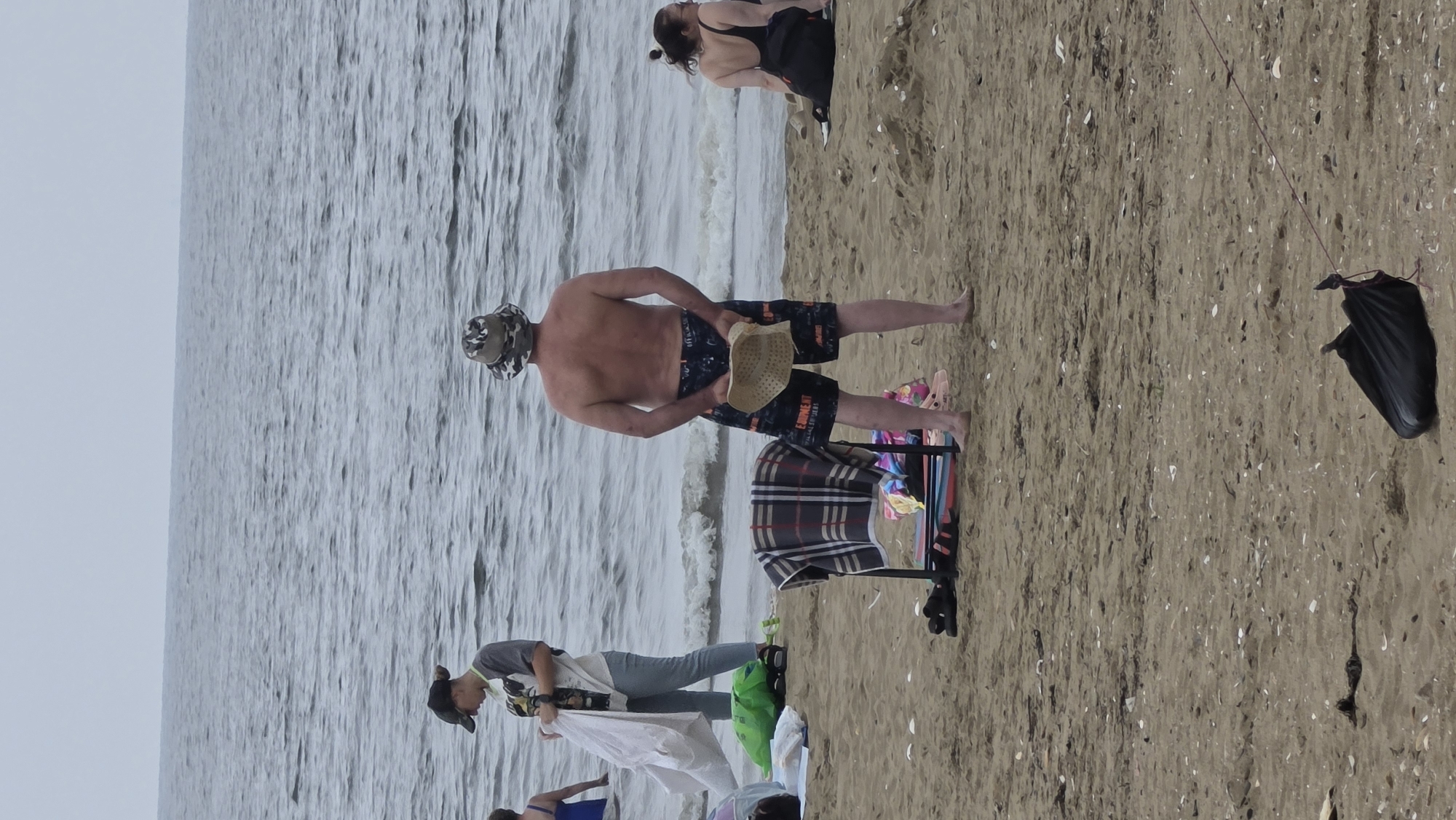 The image depicts a beach scene with several people present. In the foreground, a man wearing a camouflage bucket hat and patterned shorts stands with his back to the camera, holding a straw hat. A folding chair with a checkered blanket draped over it is positioned to his left. Further back, a woman in a dark swimsuit is seated on the sand. To the left, another person wearing a baseball cap and a t-shirt is holding a white sheet. The ocean stretches out in the background under an overcast sky. The sand is strewn with shells and debris. This scene is taking place on a beach in the city of Vladivostok, Russia, likely during the summer months given the attire of the individuals.