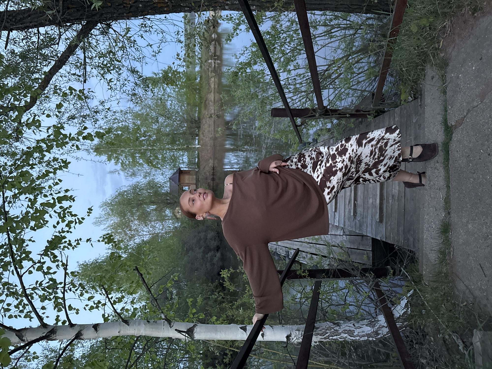 A woman stands on a wooden boardwalk near a body of water, possibly the Kama River, in what appears to be the city of Chaikovsky, Russia. She is wearing a brown long-sleeved top and a white-and-brown patterned skirt, with brown Mary Jane style shoes. She is holding onto the railing of the boardwalk. The environment includes trees, grass, and water, suggesting a natural setting integrated into the urban landscape. A building can be seen across the water. The lighting indicates it might be late afternoon or early evening.