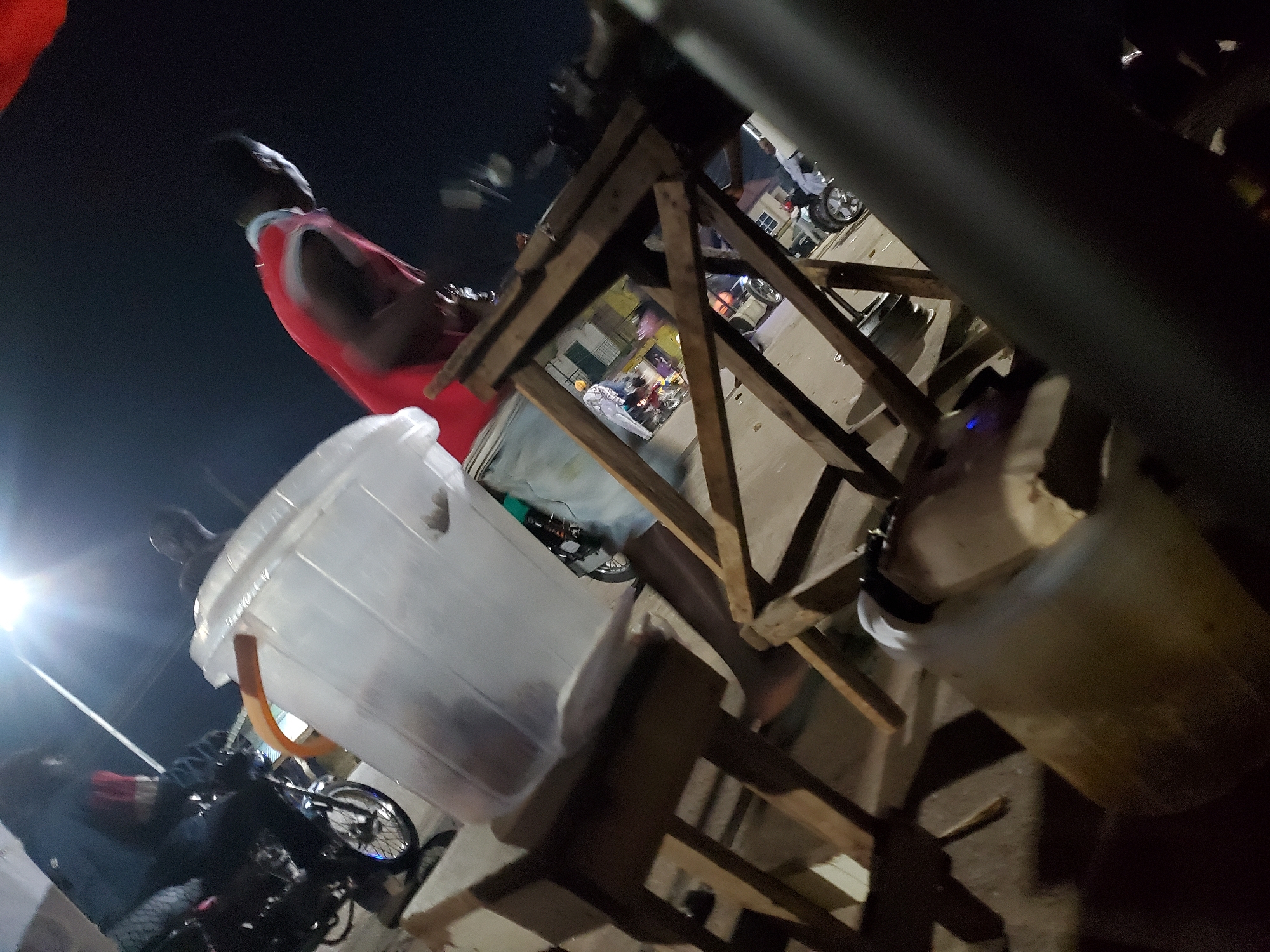 This is a nighttime, medium-angle, ground-level shot of a street scene in Gwong, Nigeria. In the foreground, a large, translucent plastic container with an orange handle sits on a wooden stool. To the left, a person is partially visible, seated on a motorcycle, wearing dark clothing and a red vest. To the right, a wooden structure, possibly a market stall, is prominent, with several people and motorcycles in the background. The lighting suggests artificial streetlights and possibly vehicle headlights illuminating the area. The overall scene depicts a bustling nocturnal environment with elements of commerce and transportation.