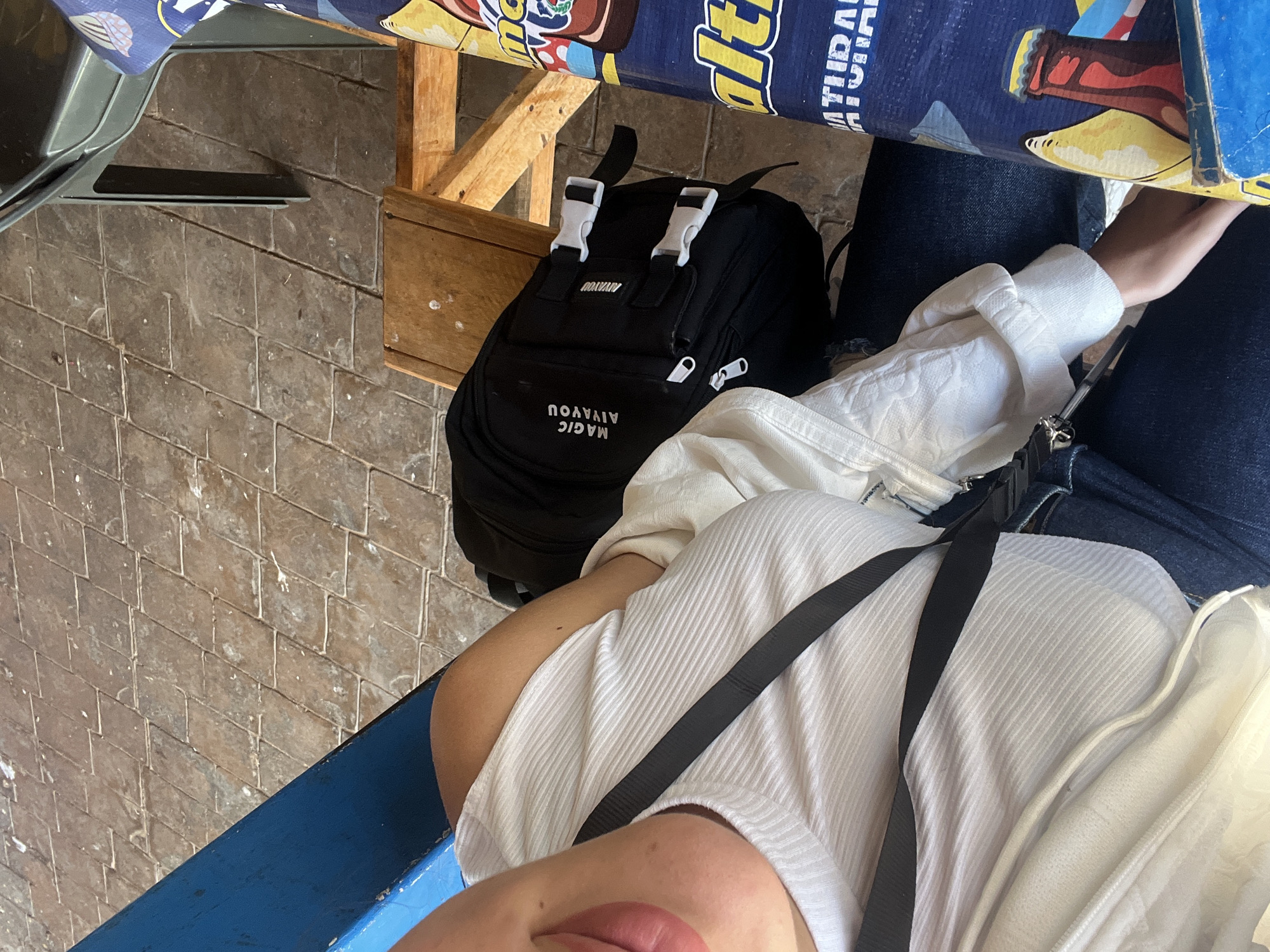 A high-angle shot captures a partial view of a person sitting at a table. The individual is wearing a white shirt with a black strap across the chest and dark-wash jeans. A black backpack labeled "MAGIC AIVAYOU" rests near the person's legs. The table is covered with a patterned tablecloth featuring images and lettering. The flooring consists of weathered, rectangular tiles. A blue painted surface is visible on the bottom left.
There is no indication of the specific location in El Valle del Espíritu Santo, Venezuela.