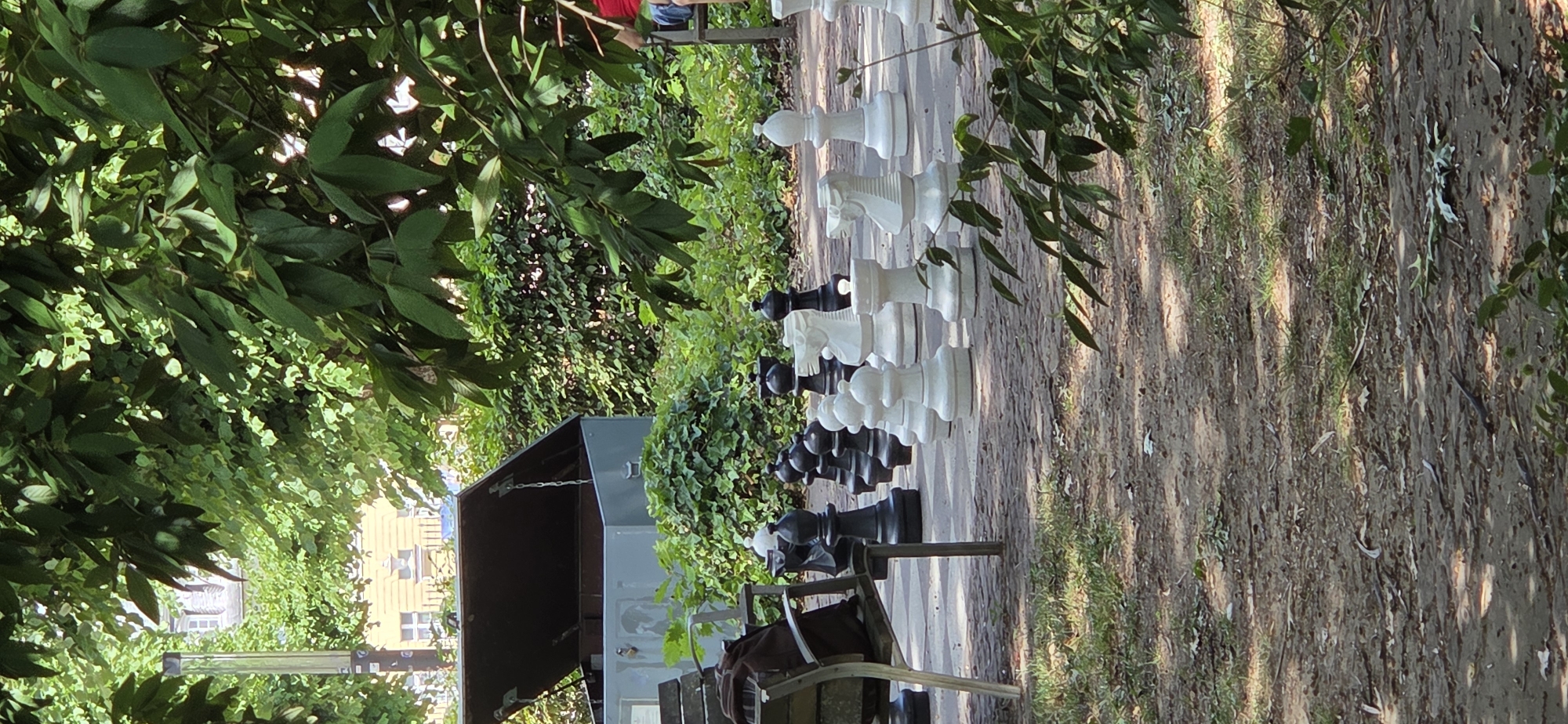 An outdoor giant chess set with large black and white pieces is positioned on a paved surface surrounded by greenery. A dark brown backpack is placed on the left armrest of a park bench, partially visible. In the background, a metal storage box with its lid open is situated amongst dense green foliage. Buildings and trees are visible in the distance.
