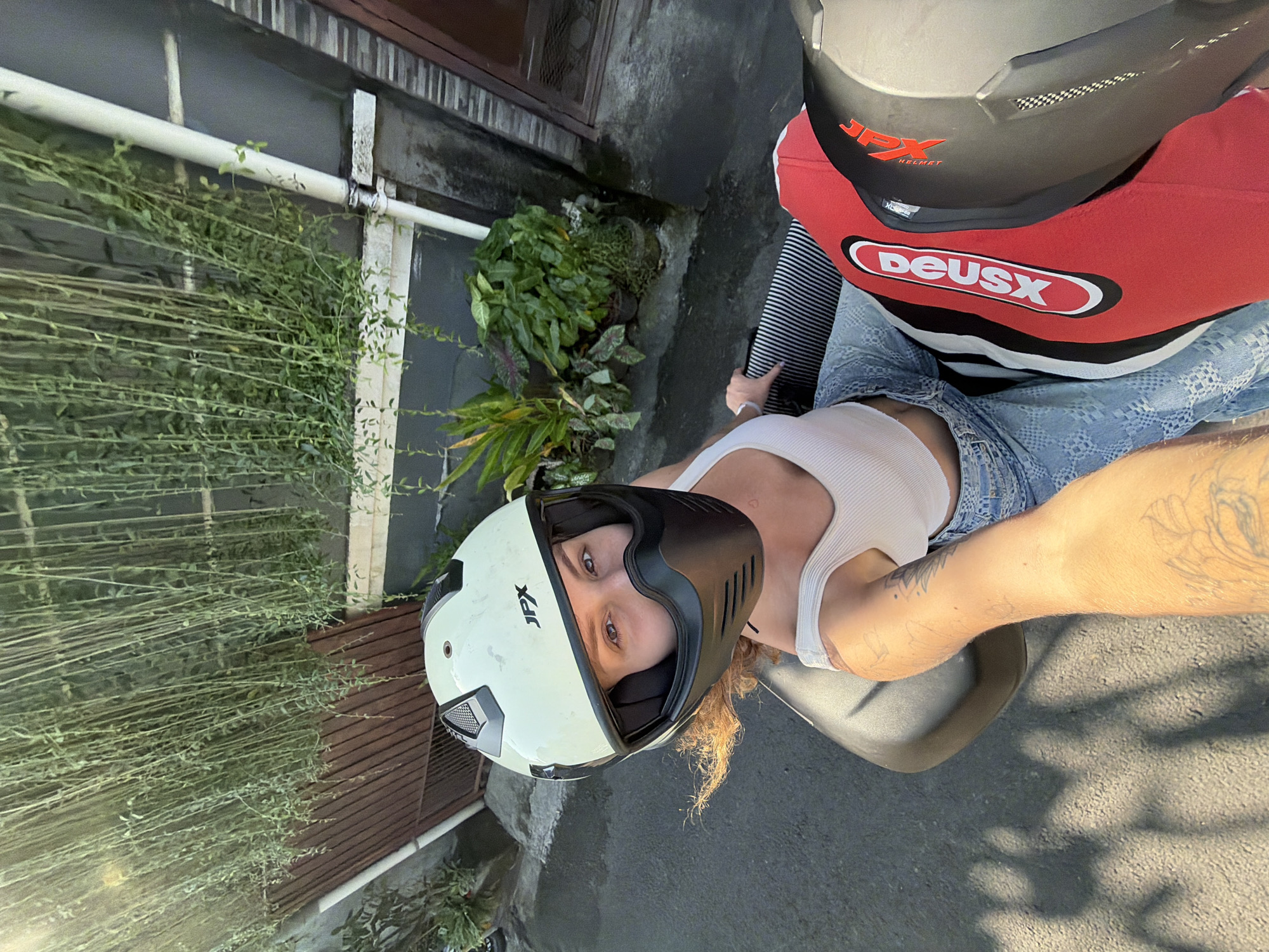 A woman with light brown curly hair, wearing a white tank top, denim jeans, and a light-colored motorcycle helmet with a black chin guard, is seated on a scooter. She is looking directly at the camera with a slight smile. Her left arm, which is extended towards the camera, displays tattoos. A second helmet, predominantly red with black and white accents, is visible on the scooter behind her. The scooter is parked on a paved surface, possibly a street, with a backdrop of lush green hanging plants and a wooden gate. The scene appears to be in a residential or urban setting in North Kuta, Indonesia, as indicated by the presence of tropical vegetation and scooter transportation. The lighting suggests it is daytime, possibly late afternoon, with the sun casting shadows on the ground.