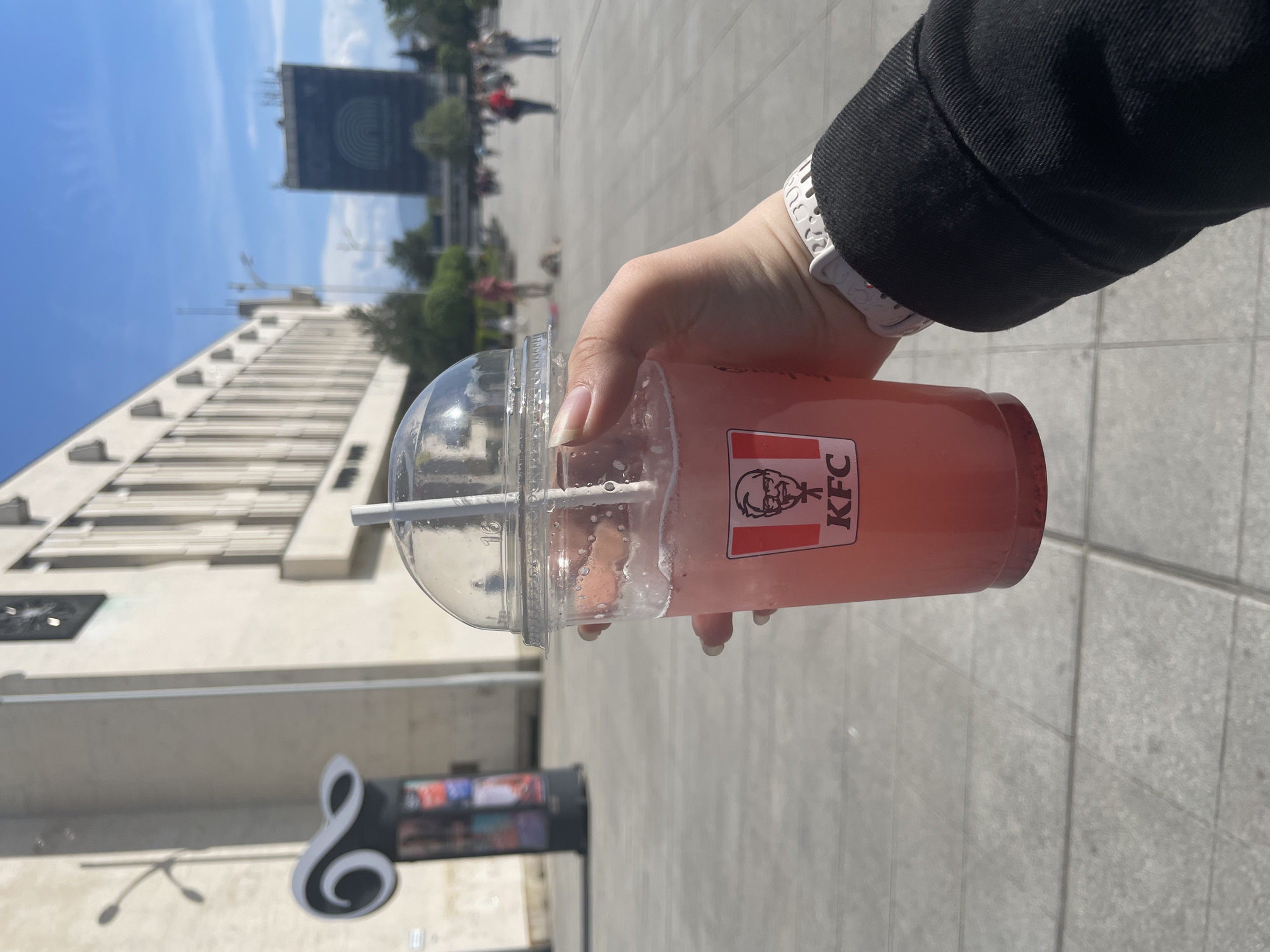 The image captures a first-person perspective of a hand holding a clear plastic KFC drink cup with a straw, filled with a pinkish-red beverage. The person is wearing a black jacket sleeve and a white, textured wristband on their left wrist. The setting is an outdoor urban plaza or square in Plovdiv, Bulgaria, bathed in bright daylight under a clear blue sky with some wispy clouds.
The ground is paved with light-colored square tiles. In the background, several buildings are visible. To the left, a large building with a modern, light-colored, ribbed facade extends upwards. Below it, a black sign shaped like a large white musical note (treble clef) is partially visible, with a colorful display underneath. Further back, a tall, dark-colored building stands out with a prominent, large, multi-colored circular or rainbow-like logo on its side. Distant, out-of-focus people and trees can be seen throughout the background, indicating a public space.
The strong sunlight creates clear shadows, suggesting a sunny day. The KFC logo, featuring Colonel Sanders' portrait and the letters "KFC", is prominently displayed on the side of the drink cup.