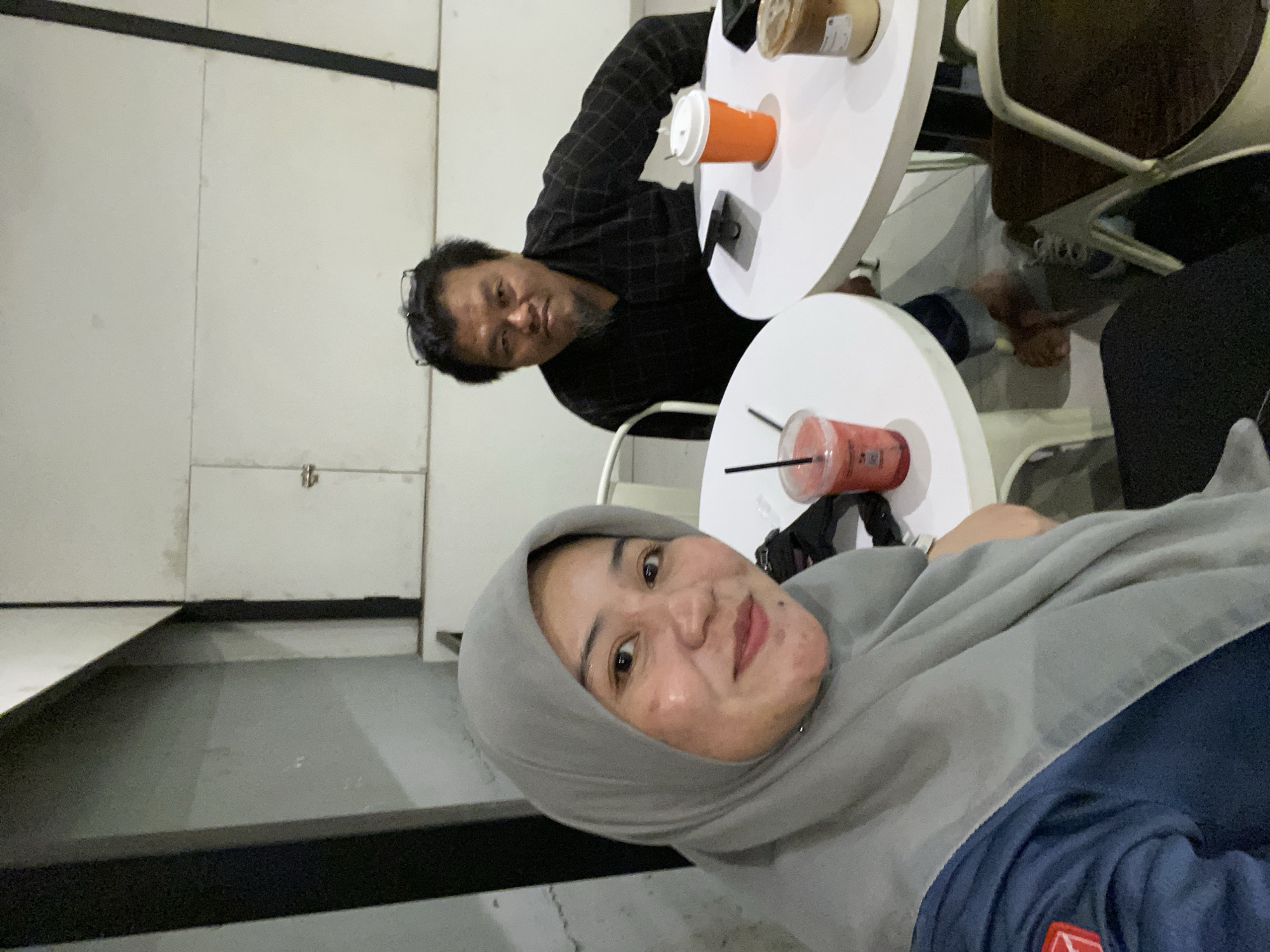 This media file shows two people, a man and a woman, at a cafe. The man is wearing a checkered shirt and glasses, and he is looking at the camera with a smile. The woman is wearing a hijab and a grey shawl, and she is also looking at the camera with a smile. They are both sitting at a white table with drinks in front of them. The man has an orange cup with a white lid, and the woman has a clear plastic cup with a red drink and a straw. The cafe appears to be indoors, with white walls and neutral-colored furniture. The lighting is dim, suggesting it might be evening or a dimly lit cafe. The people appear happy and relaxed, enjoying their time together. The image is a selfie taken by the woman, as indicated by the angle and her close proximity to the camera. No text is visible in the image. The setting suggests a casual outing at a cafe, possibly in Banjarbaru, Indonesia.