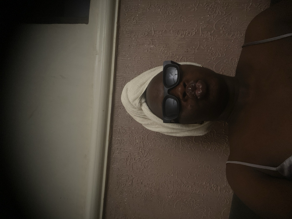 A dark-skinned person wearing black sunglasses and a cream-colored towel on their head is captured in a dimly lit room. The person's lips are pursed in a kissing expression. They are wearing a white top. The background features a patterned wall, and a white door frame is visible on the left side of the frame.