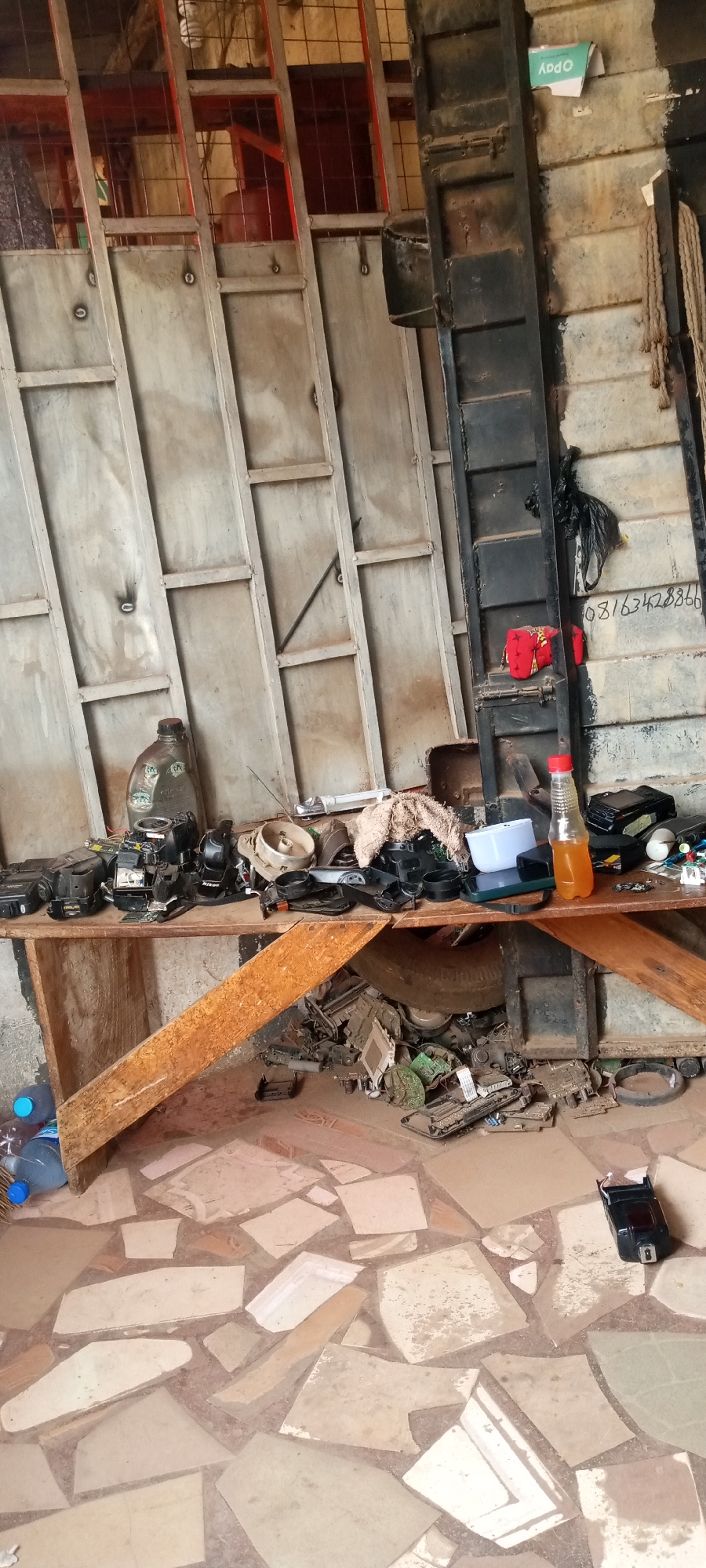 The image depicts a cluttered workspace, possibly a repair shop, located in Jos, Nigeria. A wooden table is the central focus, laden with a variety of disassembled electronic devices and components, including cameras, circuit boards, and other unidentifiable parts. A bottle of liquid, potentially juice or a cleaning solution, sits among the items. Below the table, the floor is covered in more electronic debris. The floor is tiled with mismatched stone tiles.
Behind the table, a metal wall with visible welding marks is present. A black metal ladder leans against the wall to the right. The overall impression is one of a workshop or storage area dedicated to electronic repairs. No people are visible. The environment appears utilitarian and functional, with little attention to aesthetics.