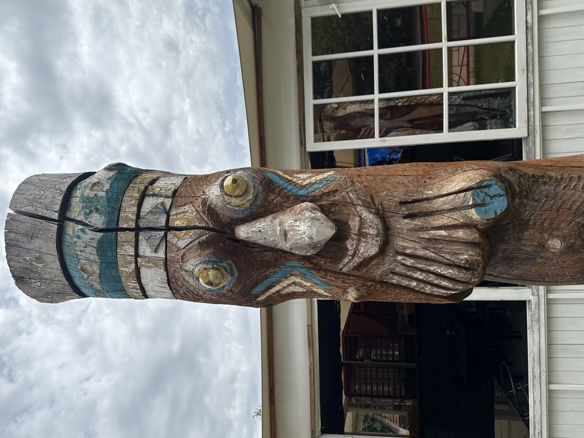 The image shows a close-up, eye-level, outdoor shot of a carved wooden totem pole with painted features. The totem pole is positioned in the foreground and occupies the majority of the frame, extending from the bottom to the top. The pole is made of weathered wood, showing cracks and peeling paint. It is decorated with carved facial features, including a nose, eyes, and a mouth, as well as geometric patterns and what appear to be stylized hands or beards. The painted colors visible are brown, blue, white, and yellow.
Behind the totem pole, there is a building with white siding and a white-framed window with multiple panes. Through the window, reflections of trees and possibly other outdoor elements can be seen. To the left of the building, a portion of another structure with an awning is visible, along with some greenery. The sky above is overcast with scattered clouds. The scene appears to be a public space, possibly an outdoor seating area or a park. The totem pole's style suggests a possible connection to indigenous art or folk art traditions.
This specific totem pole is located in the city of Yarovoye, Russia, specifically in the cultural and leisure complex "Vulkan," situated on the shores of Lake Bolshoye Yarovoye.