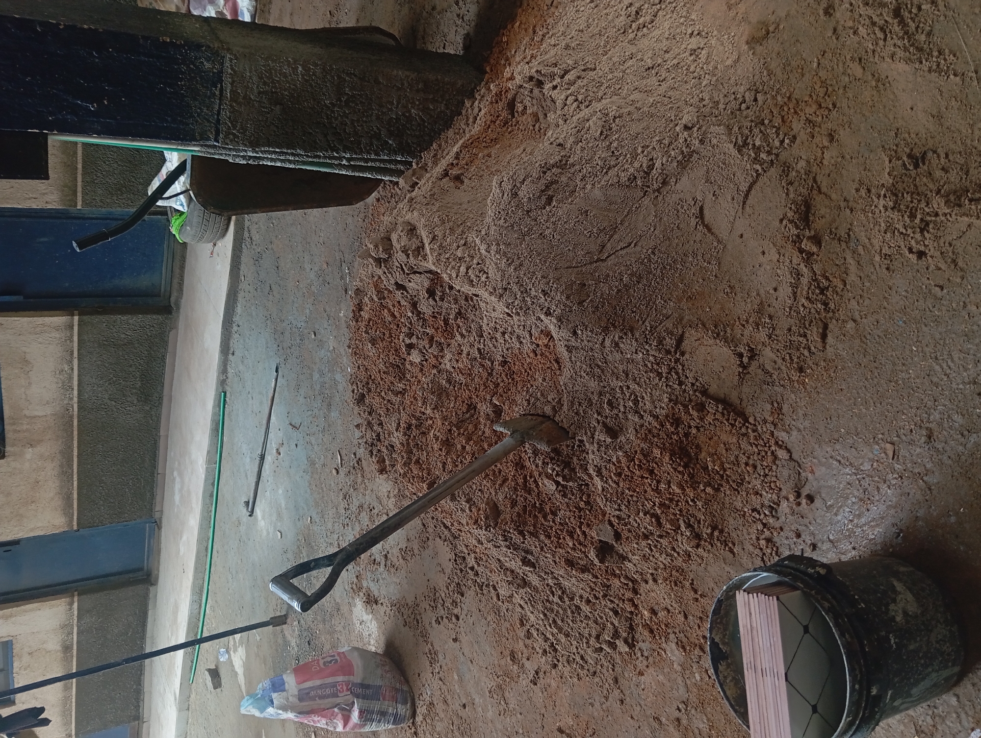 A shovel with a metal head and a black, D-shaped handle is embedded in a large pile of reddish-brown sand and dirt. To the left of the pile is a red and white bag of what appears to be cement, and to the right, a dark colored wheelbarrow with a black tire is partially visible. In the foreground to the left, a dark gray bucket contains a stack of square, light-colored tiles. The scene appears to be outdoors on a dirt or concrete surface, with a building and two blue doors in the background. A green hose is also visible on the ground.