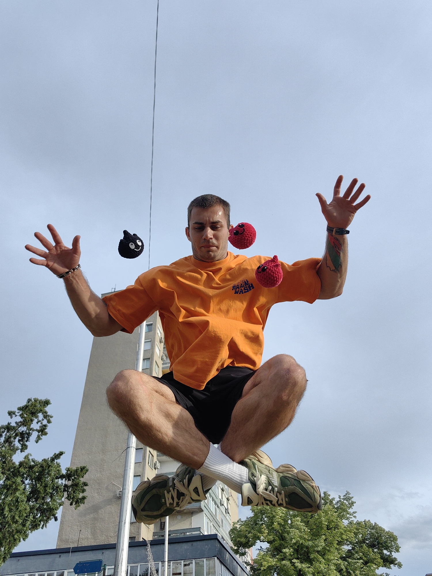 A young man in an orange t-shirt and black shorts is captured mid-air, appearing to be jumping or levitating. He is wearing white socks and camouflage sneakers. Three small, crocheted balls, two red and one black with facial features, are suspended around him, suggesting he is juggling or performing a trick. The background consists of a clear sky with scattered clouds and the facade of a modern, tall building. The lower portion of the image reveals trees and another building. The scene appears to be in an urban environment, possibly in Shumen, Bulgaria, given the architectural style of the building.