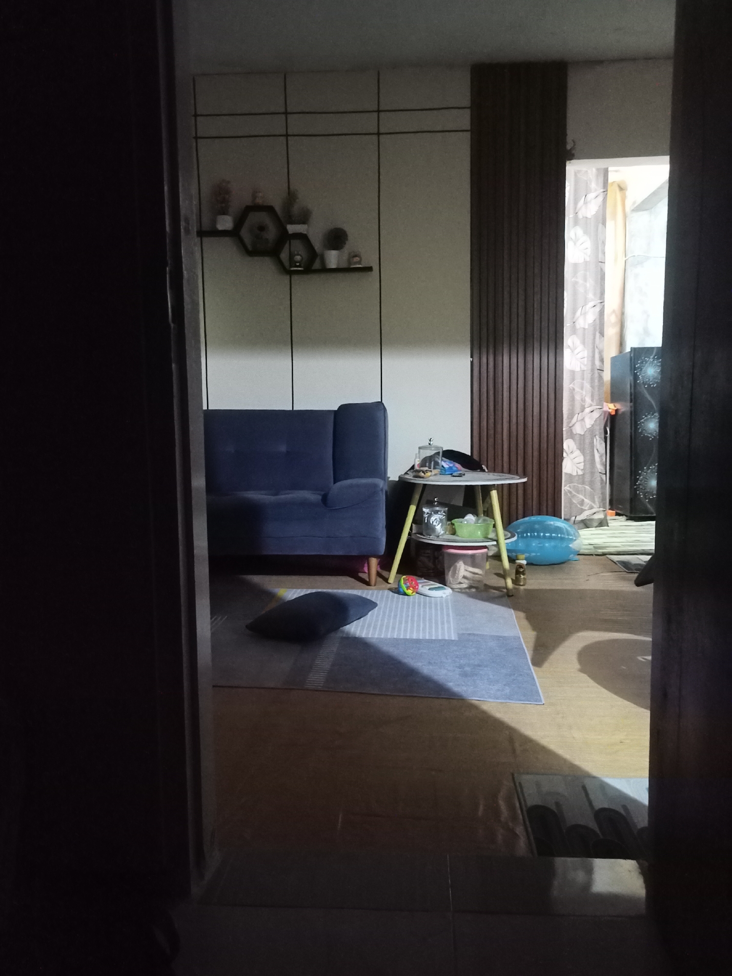 The image depicts an interior view of a living room, possibly in a residential setting in Randuagung, Indonesia. The room features a dark blue sofa, a small round side table with items on it, and a striped rug on the floor. Decorative hexagonal shelves with small potted plants are mounted on the wall. A large blue inflatable object is also visible on the floor. In the background, a doorway leads to another area with a patterned curtain and what appears to be a large electronic appliance, possibly a refrigerator. The lighting is dim, casting shadows and creating a somewhat obstructed view, suggesting it might be evening or that the curtains are drawn. The overall impression is of a lived-in space with a mix of functional and decorative elements.