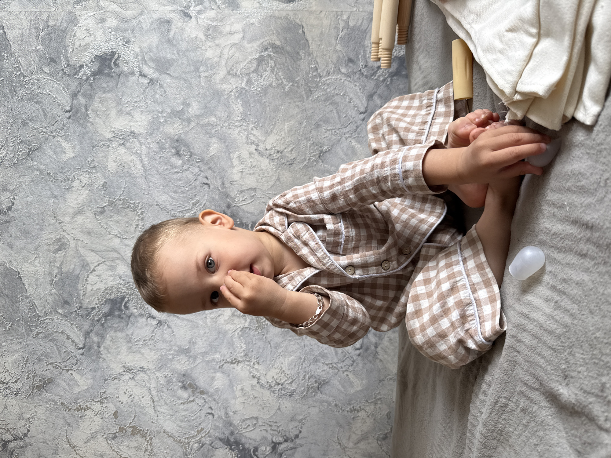 A young child wearing a brown and white checkered pajama set is sitting on a grey fabric surface, possibly a bed or couch. The child is facing the camera with their right hand near their face. They are holding a small white object near their feet. Next to the child are wooden furniture legs and white cloth. A small white plastic object is on the grey surface. The background is a wall with a textured pattern in shades of grey and white. The location is Шебунино, Russia.