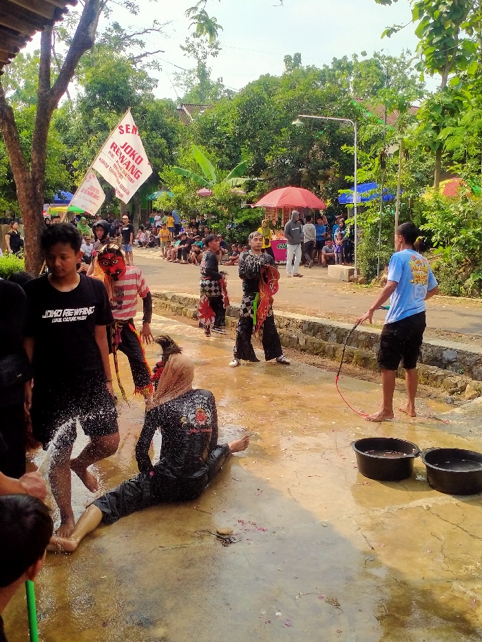 The scene captures an outdoor event in Gebyog, Indonesia. In the foreground, a person in black long-sleeved attire with a light-colored head covering is seated on a wet, light-colored paved surface, being actively splashed with water. Red flower petals are scattered in the water around this individual. To their left, another male individual in a black T-shirt and dark shorts, barefoot, stands in the immediate vicinity of the splashing water. In the mid-ground, a person wearing a red mask with white teeth and a striped shirt stands on the wet ground, alongside two individuals dressed in traditional Indonesian dance costumes featuring dark pants and patterned tops. Further to the right, a barefoot male in a light blue T-shirt and dark shorts holds a red-tipped whip or rope, positioned near two black plastic buckets filled with water. The background features a crowd of spectators, many seated along a raised concrete embankment, observing the event. A paved road or path is visible behind the performers, leading to vendor stalls under a red umbrella and a blue canopy. The overall environment is lush with numerous trees and green foliage under a clear, bright sky.