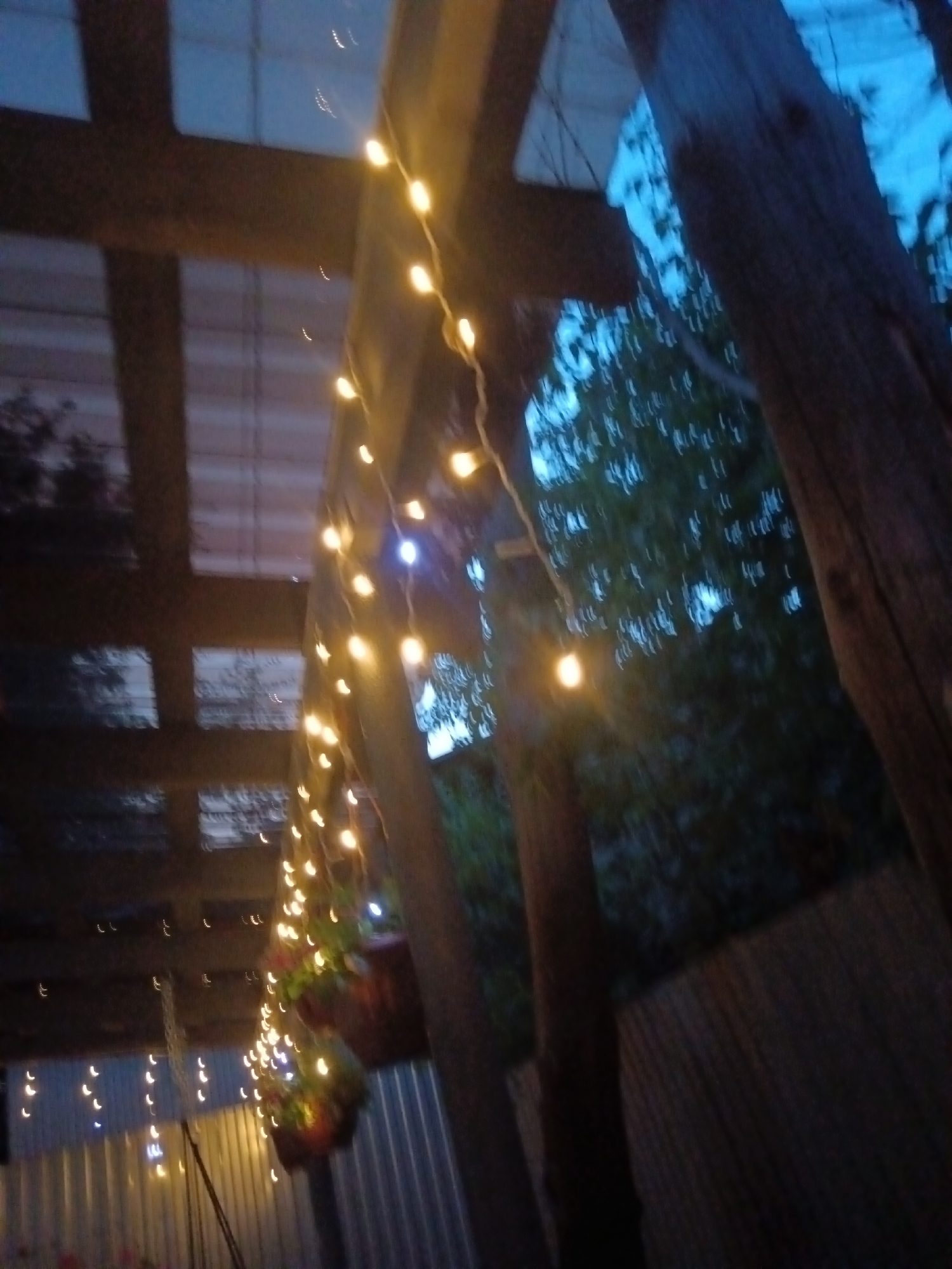 The image depicts an outdoor scene at dusk, likely in Sorochinsk, Russia. The foreground is dominated by several wooden posts and beams supporting a translucent roof structure, possibly part of a veranda or pergola. A string of warm-toned fairy lights is draped across this structure, illuminating the scene with a soft glow. In the background, dark green foliage is visible, interspersed with areas of blue sky, suggesting the presence of trees and indicating the time of day. The overall atmosphere is subdued and perhaps a bit rustic, with the hanging lights adding a touch of decorative ambiance. No people are present in the image.