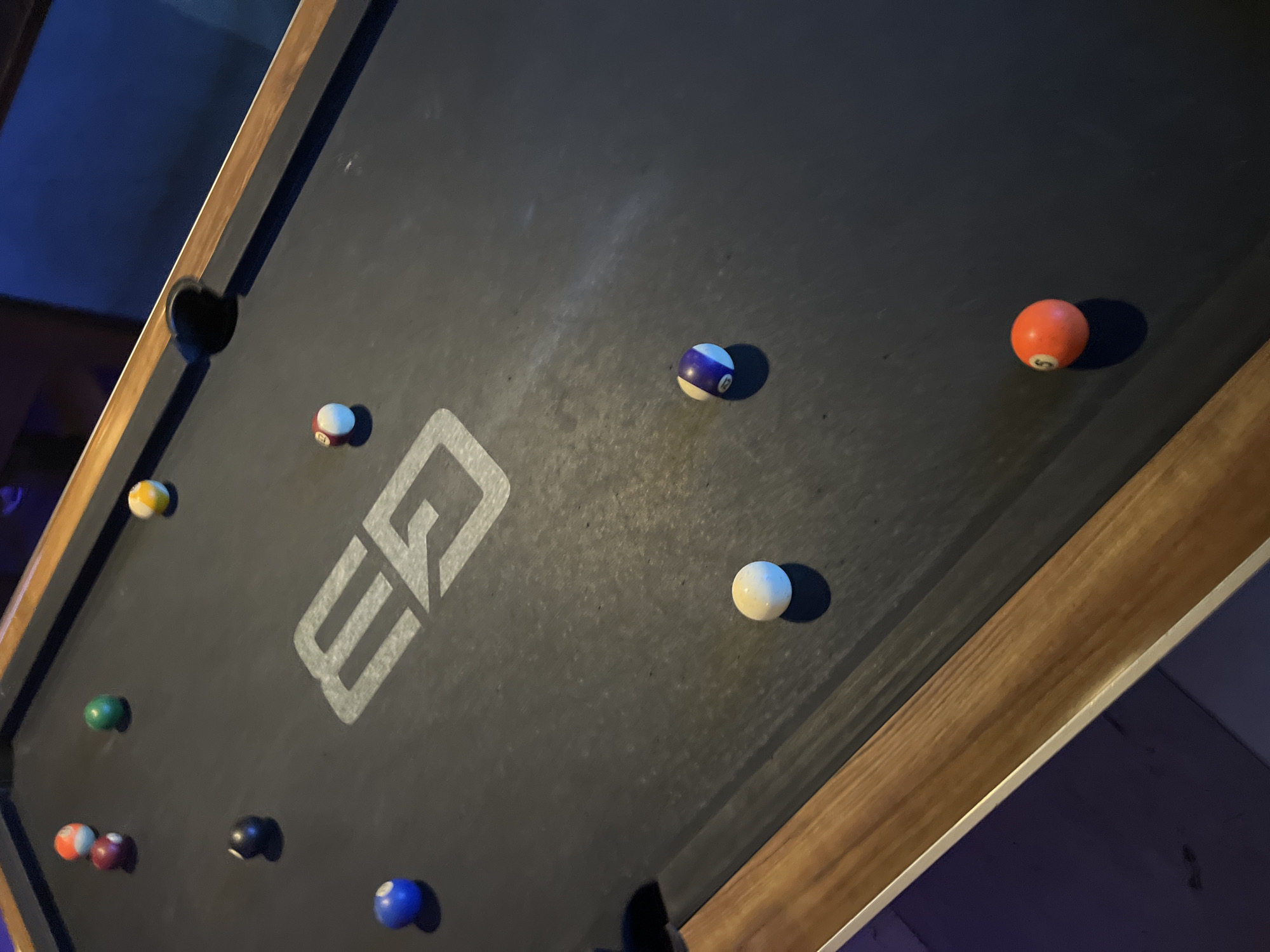 A dim, angled photograph captures a pool table with a dark felt surface, illuminated by low artificial light, hinting at an evening or nighttime setting in an indoor venue such as a bar or game room. The table features a rich wooden frame with a distinct metallic, possibly silver, trim. One black pocket is visible in the upper left corner of the frame.
Several billiard balls are scattered across the table, suggesting a game of pool is either in progress or has recently concluded. Notable balls include a bright orange "5" ball in the upper right, a blue and white striped ball near the top center, and a white cue ball positioned mid-right. Towards the bottom left, a green solid ball, a red and white striped "1" ball, and a yellow and white striped ball are visible. A dark solid ball (possibly the 8-ball) and a blue solid ball are also present towards the bottom of the frame.
A prominent white "EQ" logo is centered on the dark felt of the pool table. Apart from the numbers "5" and "1" on their respective balls, no other distinct text is clearly legible. The overall ambiance is relaxed and recreational, typical of a billiards hall. The scene is devoid of people, focusing solely on the game setup. The blue hue reflecting in the background suggests ambient lighting within the space. While the location context of Barcelona, Spain, is provided, there are no specific visual cues within the image itself to confirm this.