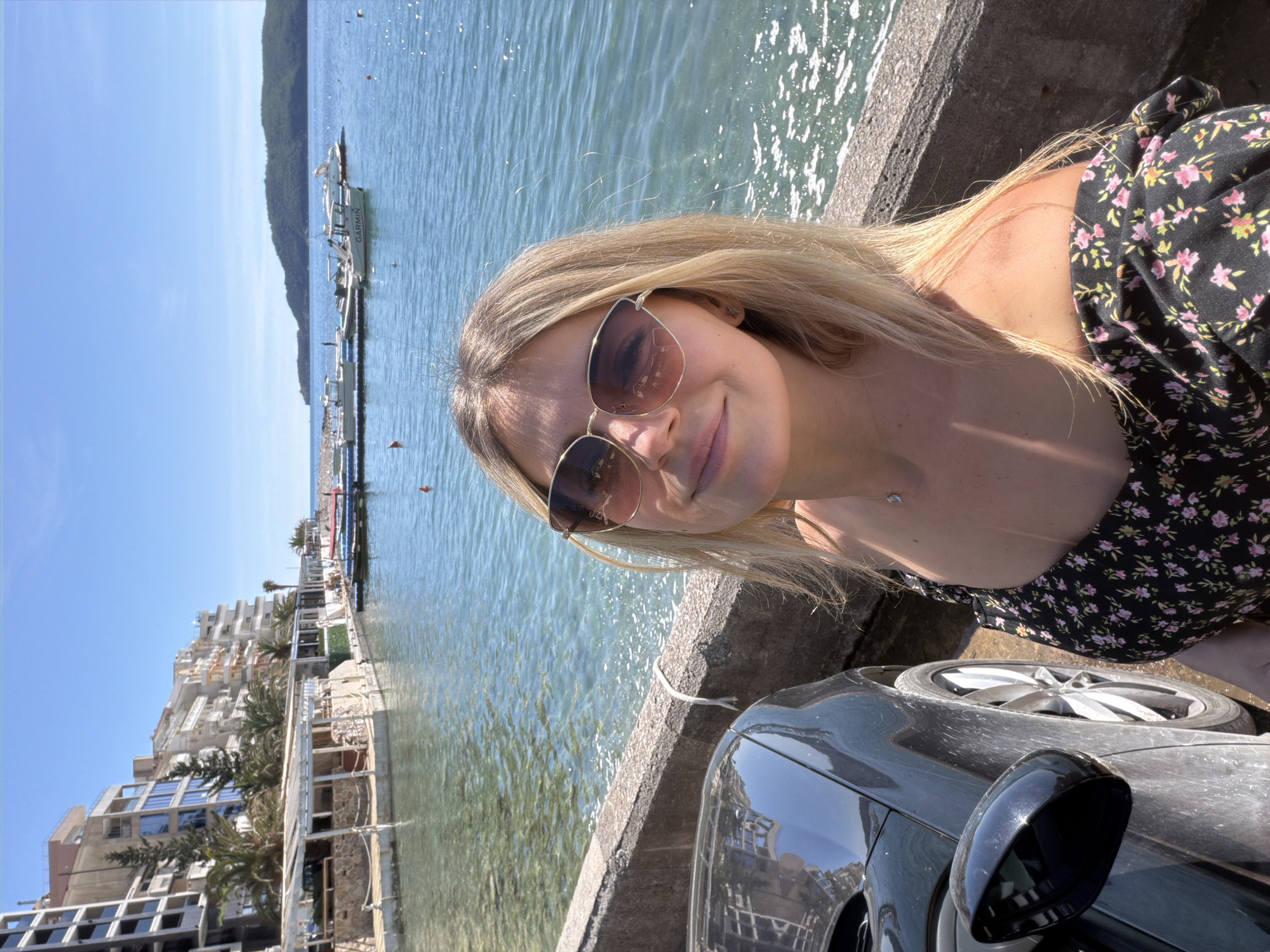 A medium-shot selfie of a blonde woman with sunglasses on a concrete wall overlooking the Adriatic Sea in Rafailovići, Montenegro. Buildings of a hotel complex are on the left and a marina with boats is in the background. A dark gray car is in the foreground on the right. The sky is bright blue.