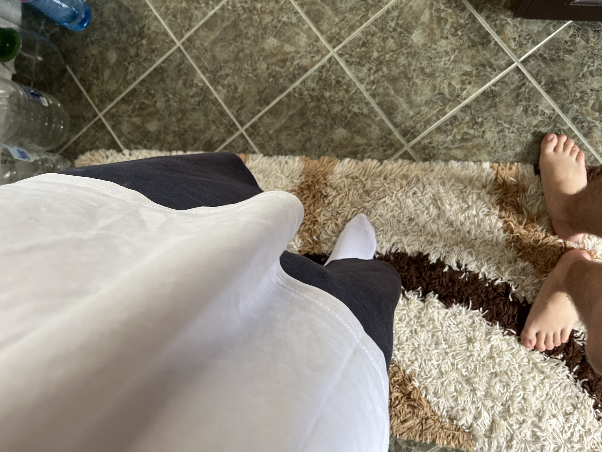 An eye-level, indoor photograph shows a person sitting on a patterned rug in what appears to be a home. The person wears black pants, a white sock on their left foot, and a large white shirt. The person's right foot is bare. The rug has a brown and white striped pattern. The floor is tiled with a dark brown geometric pattern. In the top left of the image, a stack of water bottles is visible. The location is Zlatograd, Bulgaria.