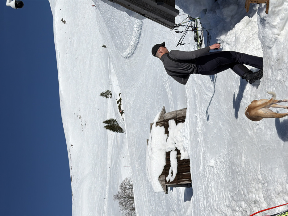 The image depicts a bright, snowy mountain scene in Vranjak, Montenegro. In the foreground, a male individual wearing a dark long-sleeved turtleneck shirt, dark pants, and a dark baseball cap walks through deep snow, facing away from the camera and slightly to the right. A light brown and white dog follows closely behind him, seen from the rear. To the man's immediate right is a partial view of a wooden structure, against which several pairs of skis and poles are propped. A white dome-shaped surveillance camera is mounted high above this structure, oriented towards the distant ski slopes. To the left of the man and dog, a rustic wooden shack is partially visible, its roof and front heavily covered in snow. The midground and background consist of extensive snow-covered ski slopes exhibiting numerous ski tracks. Several evergreen and bare deciduous trees are scattered across the slopes. Multiple small figures, identifiable as skiers, are visible at various points on the distant slopes. The sky is clear and deep blue, and bright sunlight casts sharp shadows across the snow.