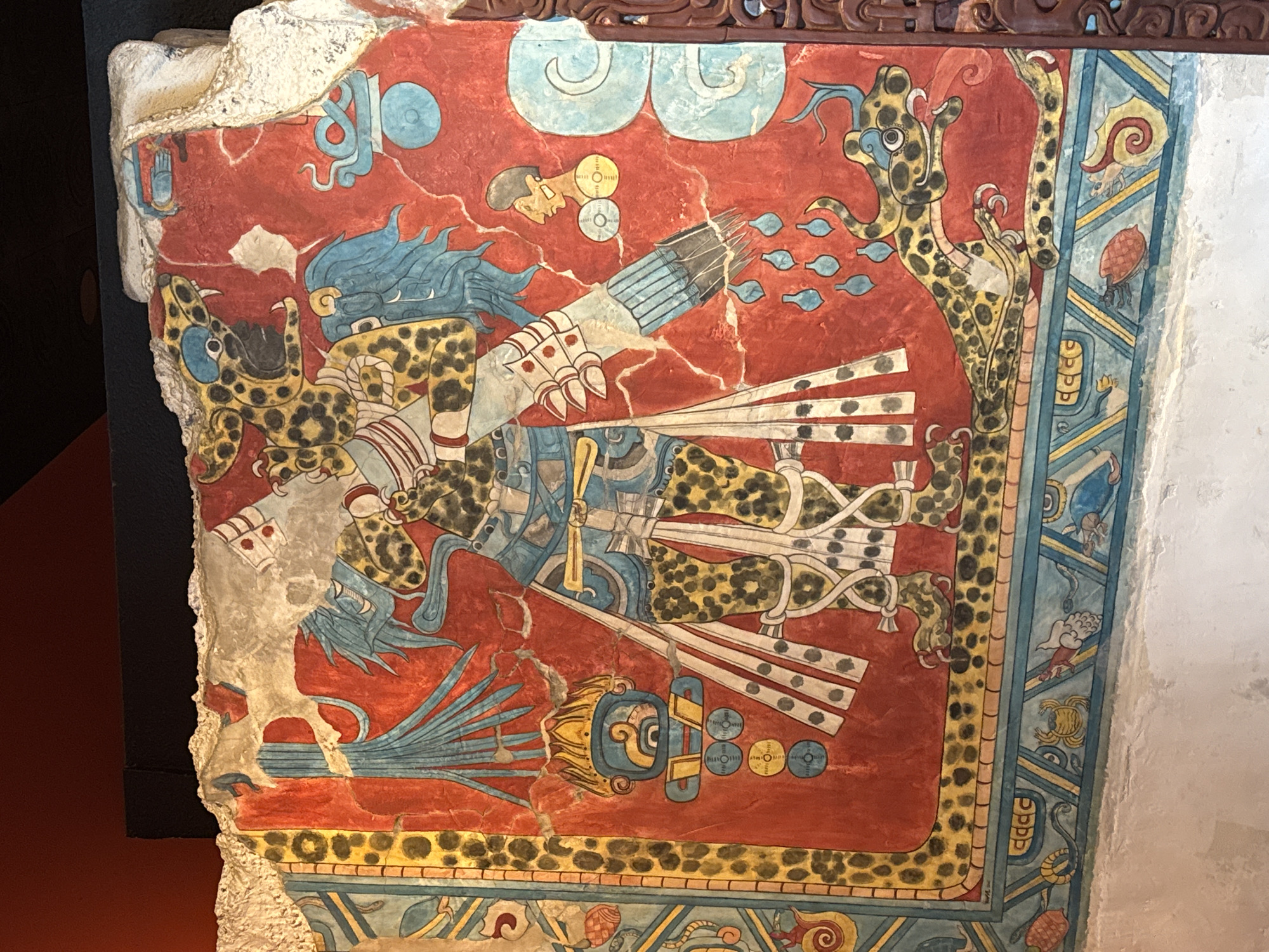 The image depicts a fragment of a painted mural, likely a segment of a larger composition. The mural fragment features a vibrant, predominantly red background with representations of figures and symbolic elements rendered in a stylized, ancient Mesoamerican artistic tradition. The main figure appears to be a jaguar-like character, possibly a deity or a high-ranking individual adorned in jaguar skin regalia. The figure is depicted in profile, with blue flowing hair or headdress elements. The mural also includes depictions of spears or arrows, and other geometric and representational forms such as clouds and an abstract portrait. The fragment is bordered with a frame containing a variety of oceanic creatures: snails, crabs, shellfish, and eels.
The artifact is likely located in a museum or historical site in Mexico City, Mexico, as it is indicative of the ancient Mesoamerican cultures that thrived in that region. The artifact appears to be affixed to a light colored wall with modern infrastructure present.