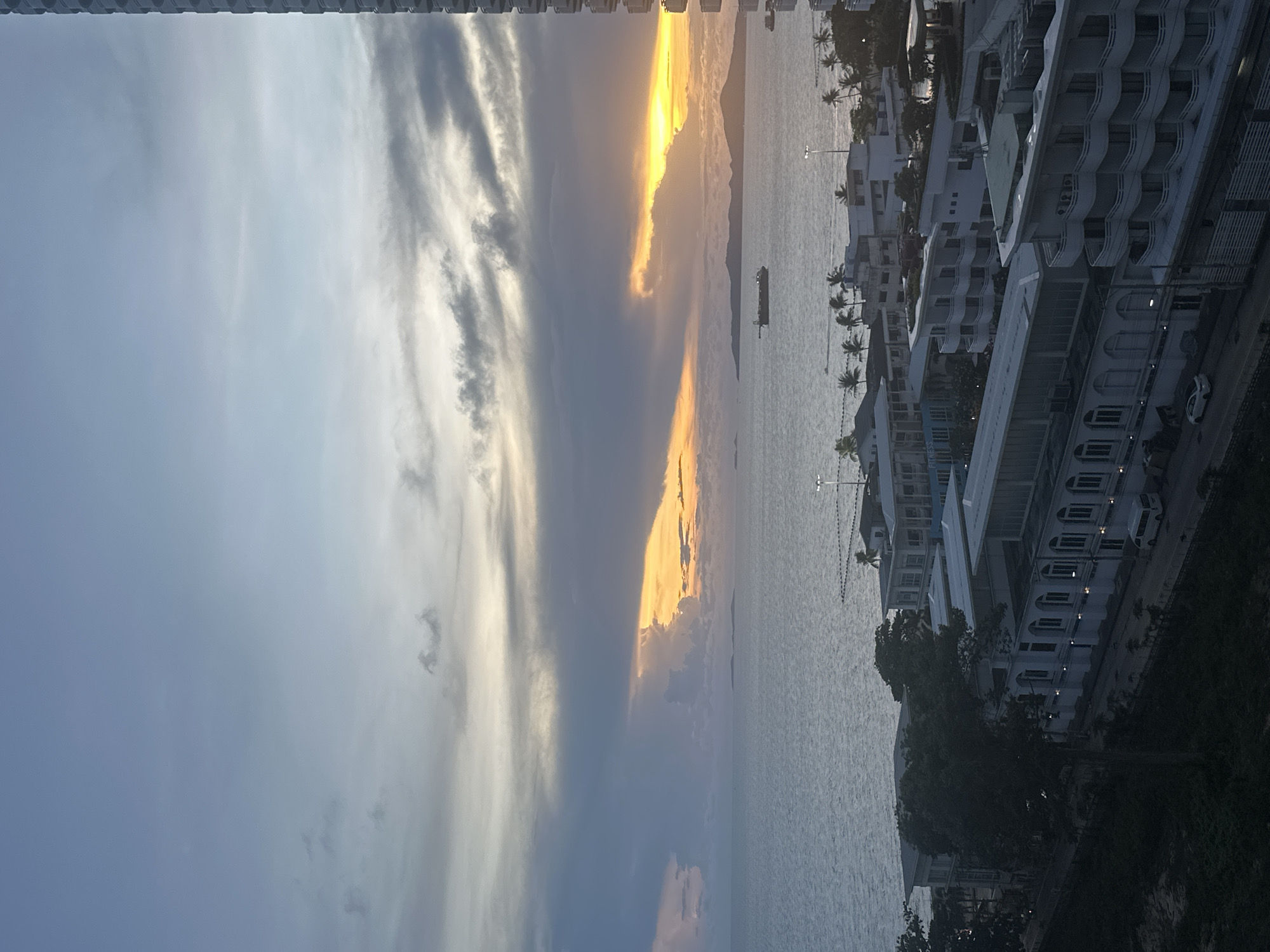 The photograph depicts a coastal scene in Pattaya City, Thailand, taken during sunset. The sky is partially obscured by clouds, with a band of golden sunlight illuminating the horizon and reflecting on the water. A cargo ship is seen in the distance. On the right side of the frame, multiple white buildings with balconies are visible, interspersed with palm trees. Streetlights line the coast, and a road with parked cars can be seen adjacent to the buildings. The overall scene suggests a view from an elevated position, likely a high-rise building.