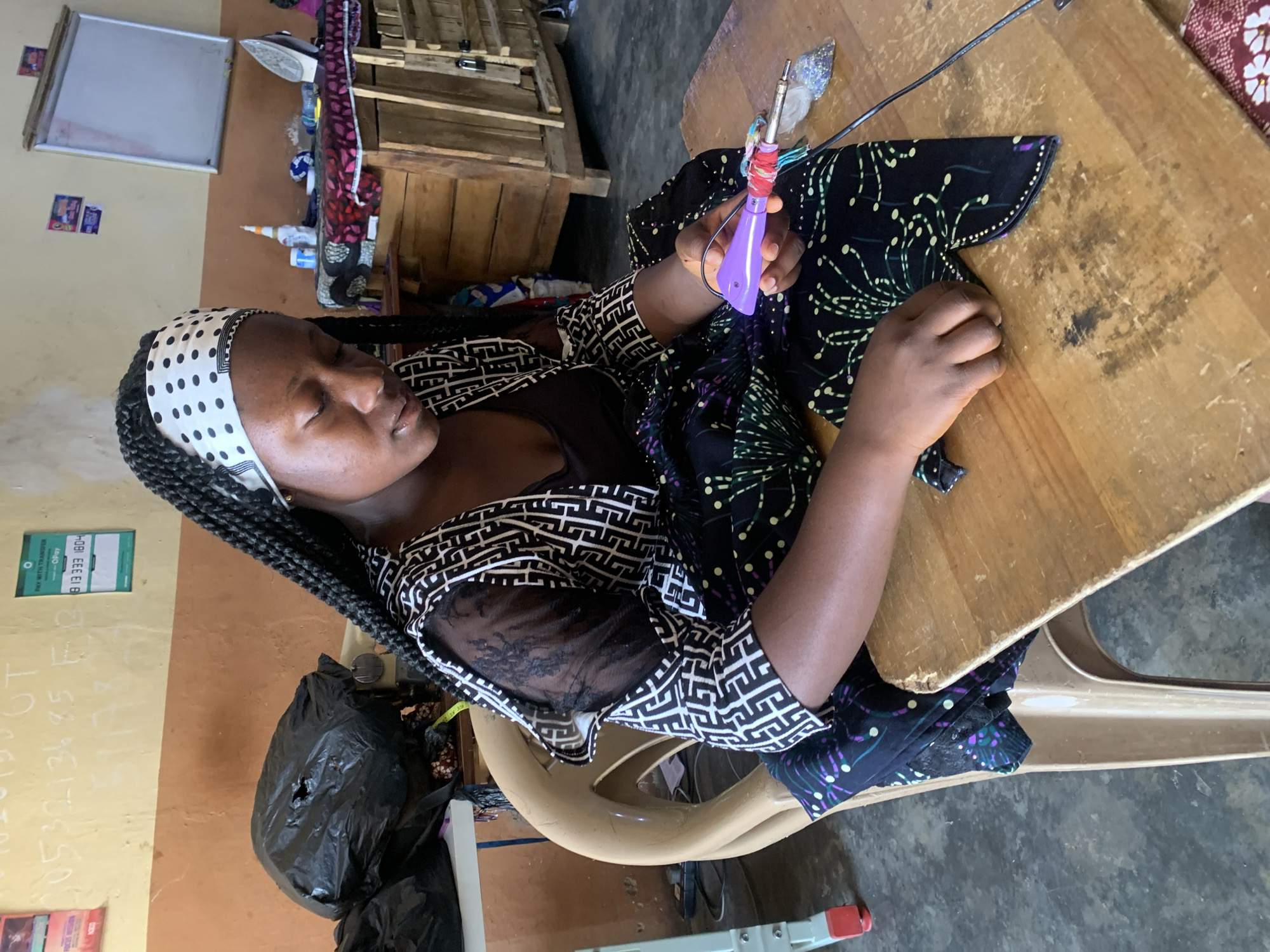 A woman is seated at a wooden table, seemingly working on a sewing project. She is using an electric tool with a purple handle on a piece of patterned fabric. The woman is wearing a black and white patterned shirt, a black top, and a white headscarf with black polka dots.
The room appears to be a work area, possibly a small business. A black trash bag is visible near her chair, and numbers and text are written on the wall. A sign that reads “PAY WITH TRANSFER OPAY” is also visible on the wall with the phone number 813 333 1804. A wooden cabinet sits in the background.
Based on the sign on the wall and the cultural context suggested by the woman's attire and the patterned fabric, the location is likely Jos, Nigeria.