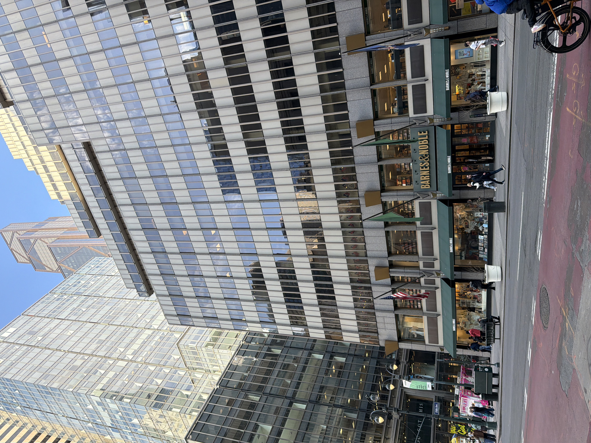 Here is a description of the image:
The image shows a street scene in New York City, United States of America. A Barnes & Noble bookstore is visible along the right side of the image, indicated by its signage. Pedestrians are walking on the sidewalk and crossing the street. The street itself is paved with red lanes. Additionally, street lamps are seen lining the sidewalk near the buildings. Tall skyscrapers dominate the background, featuring a grid-like window pattern. The sky is partly visible.