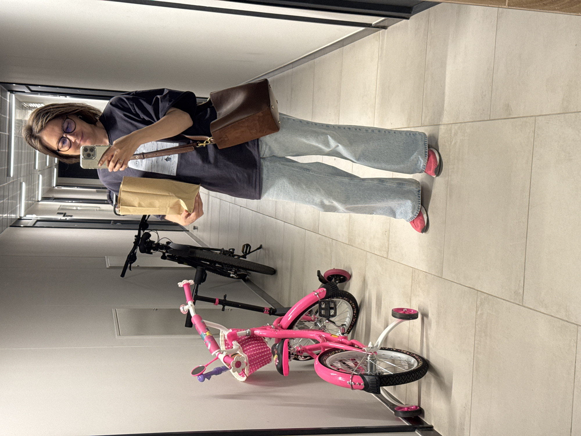 A woman wearing glasses and a dark t-shirt, holding a brown paper bag and a mobile phone, stands in a well-lit apartment building hallway in Izhevsk, Russia. She is also carrying a brown crossbody bag. In front of her, leaning against the wall, are two bicycles: a small pink children's bicycle with training wheels and a basket, and a larger black bicycle. The hallway has tiled floors and plain white walls with some light fixtures visible on the ceiling.