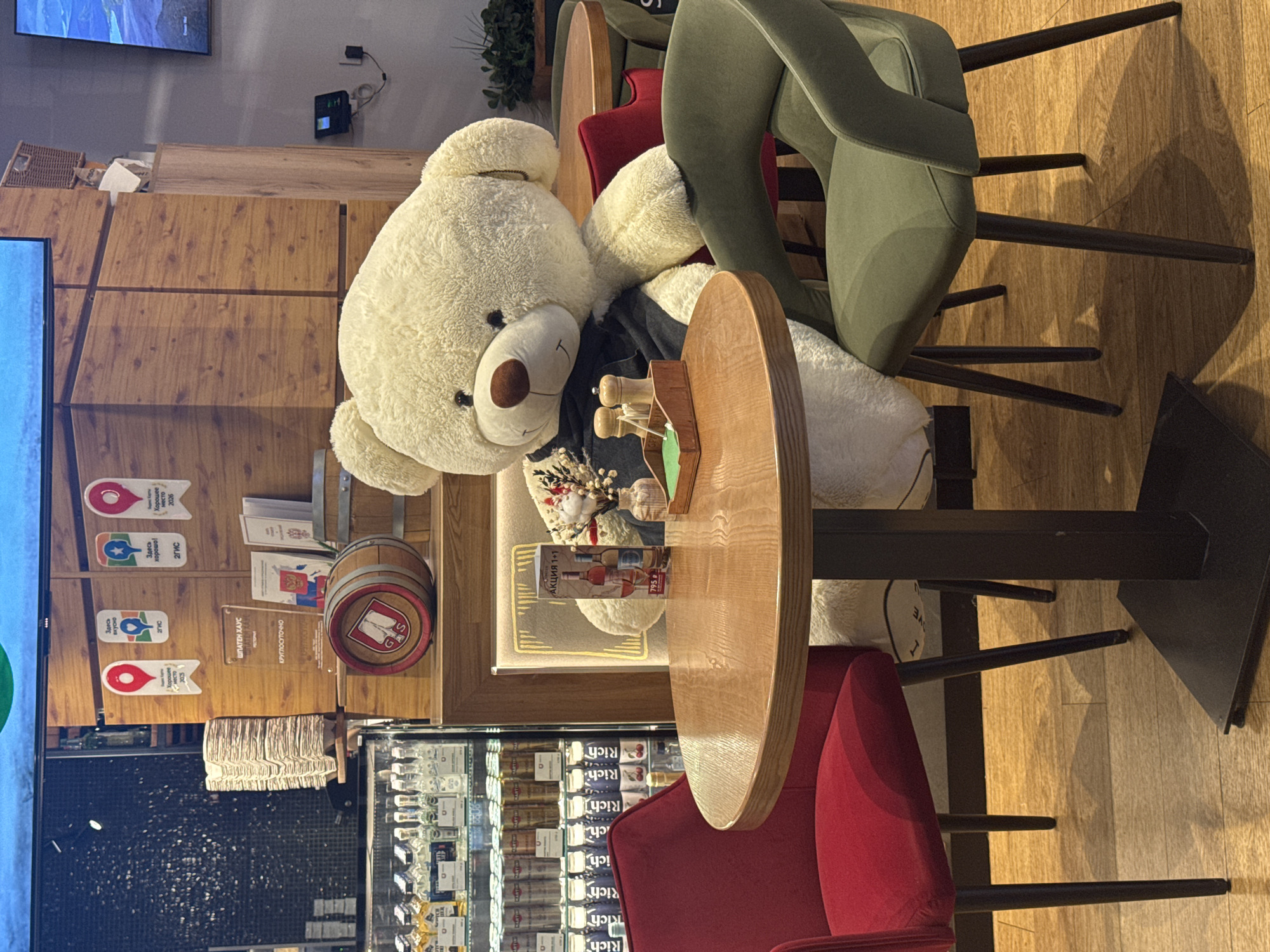 The image shows the interior of a restaurant or bar in Sochi, Russia. A large, white teddy bear sits at a small, round wooden table, partially obscuring the view. On the table, there are various objects including a small wooden display with toy figures, a bouquet of flowers, and a promotional card displaying the text "АКЦИЯ 1+1" and "795" in a foreign language. Behind the teddy bear, on the wall, are various plaques and decorative items, including a miniature barrel. A sign with Cyrillic text, "ШПАТЕН ХАУС КРУГЛОСУТОЧНО" (potentially indicating the establishment's name and operating hours), along with smaller plaques with the text "Здесь хорошо! 2ГИС" and "Здесь вкусно 2ГИС" are also visible. The seating consists of a green velvet chair and a red chair, indicating a seating arrangement around the table. In the background, a refrigerated display case is filled with beverages labeled "Rich." The image captures a unique and whimsical atmosphere within the establishment.