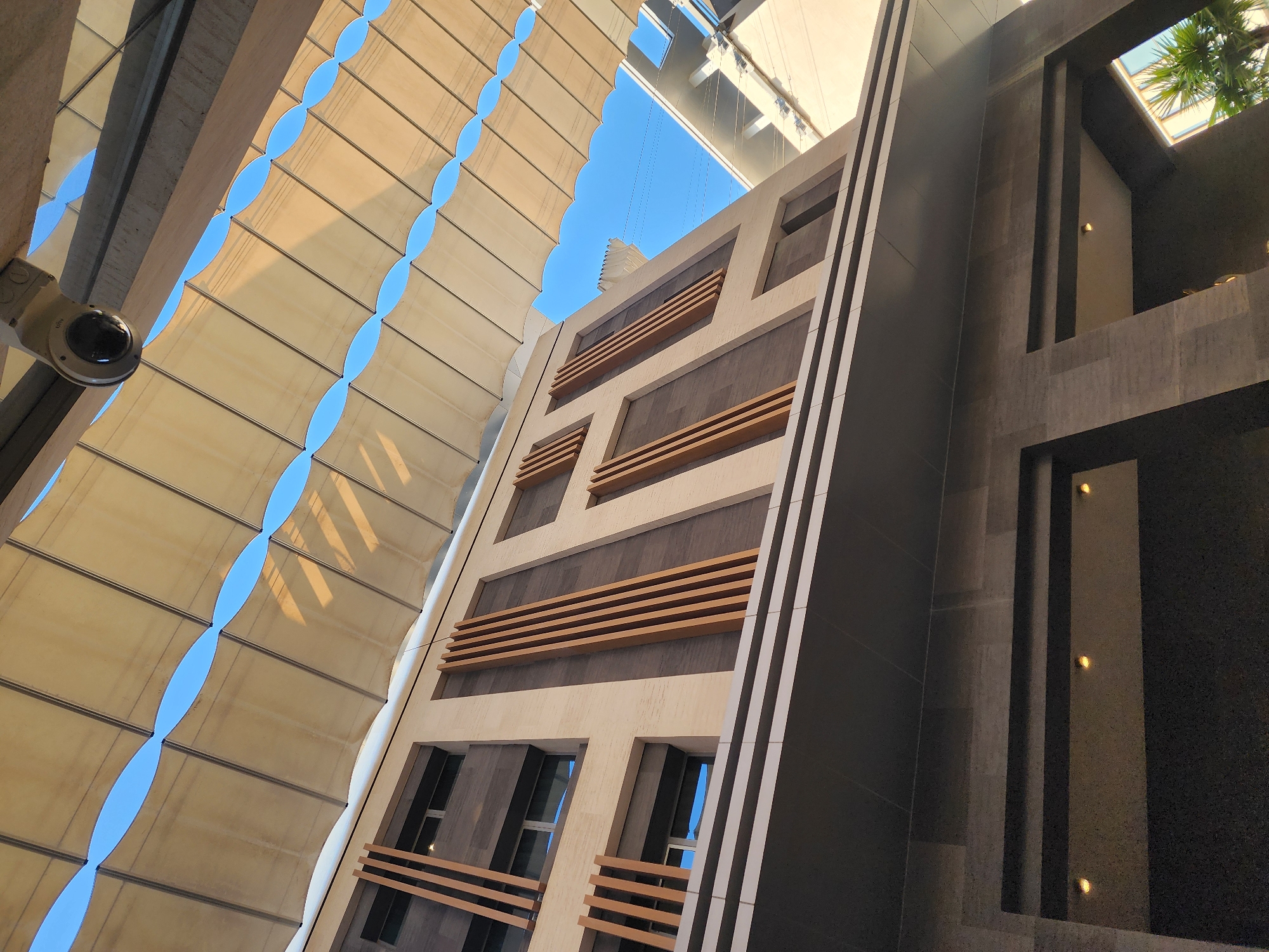 The image shows the exterior of a modern building with a distinctive facade, located in Al Rayyan, Qatar. The building features multiple floors with a combination of light-colored stone and dark paneling. Vertical wooden slats are a prominent design element on the upper floors, interspersed with rectangular windows.
Above the building, a large, light-colored fabric canopy stretches across, creating a series of oval openings that reveal a clear blue sky. Sunlight casts shadows from the canopy onto the building's facade, creating a dynamic interplay of light and shade. A security camera is mounted on an overhead beam, directing its lens downwards towards the area below. The lower part of the image shows an overhang with dark paneling and recessed lighting. The overall impression is one of contemporary architecture in an urban setting, possibly a commercial or public space.