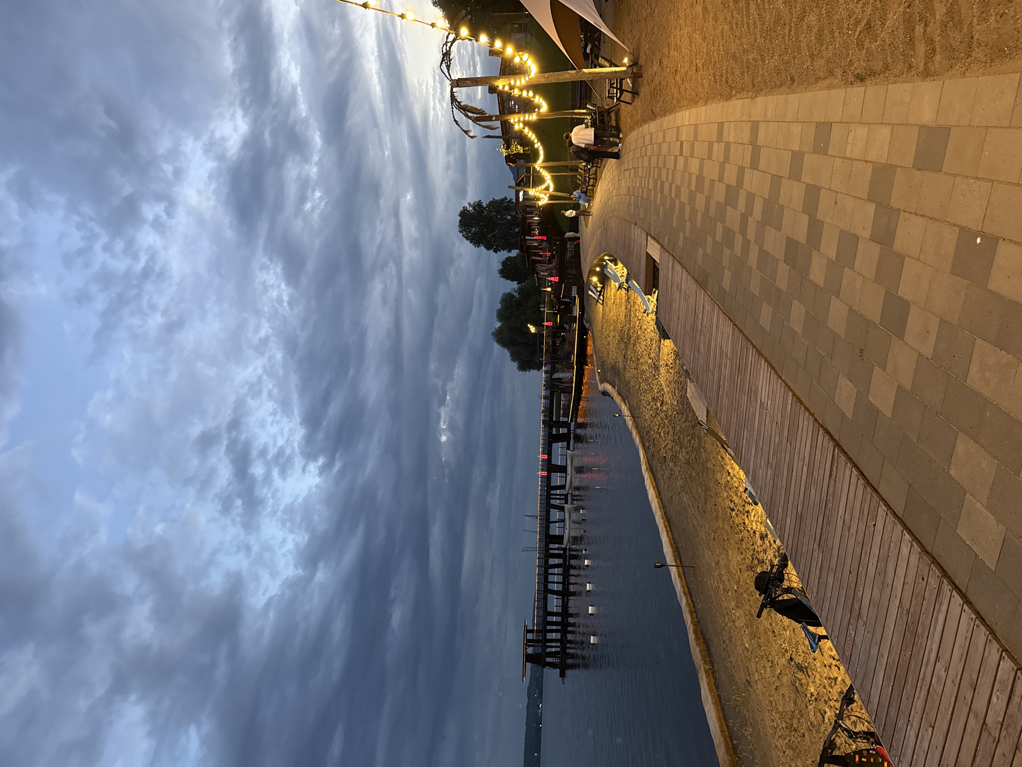 This is a vertical, eye-level, medium-shot photograph of a waterfront walkway in Votkinsk, Russia, at dusk. The sky is filled with dramatic, dark clouds, with patches of lighter blue visible. A calm body of water, likely the Kama River, extends across the left side of the frame. On the right side of the water is a sandy beach, bordered by a wooden boardwalk. Further to the right, a paved walkway with checkered pattern tiling curves into the distance. String lights with glowing yellow bulbs are draped across poles and palm trees, illuminating the walkway and nearby seating areas. Several people are visible, some standing and talking, others sitting at tables. A long pier extends into the water on the left, featuring a covered structure at the end and red lights along its length. A small white boat is docked near the pier. Two bicycles are parked on the wooden boardwalk in the lower left corner. The overall scene suggests a relaxed evening atmosphere at a waterfront establishment.