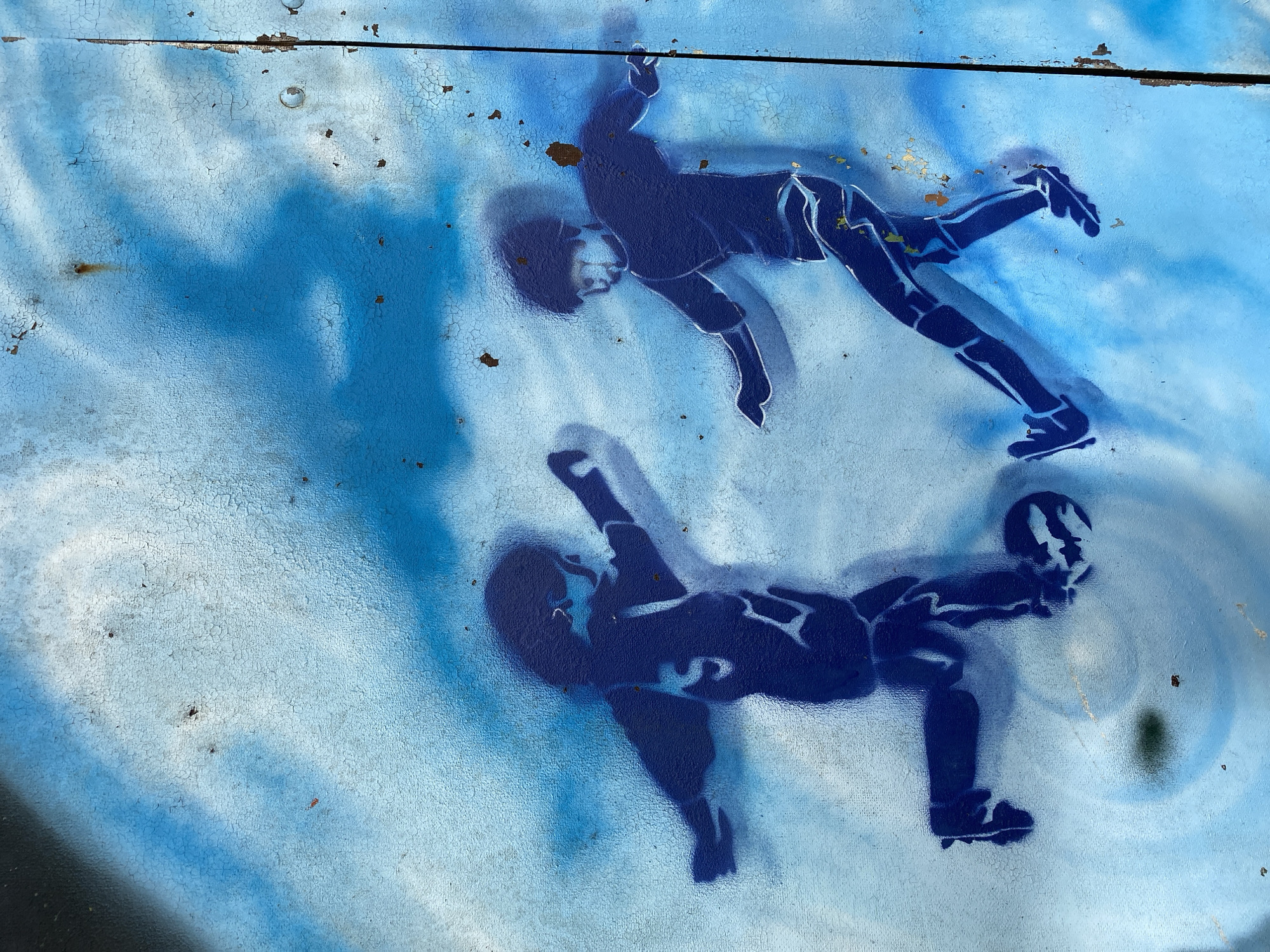 The image depicts a stencil artwork on a light blue, cracked, and weathered surface, possibly a metal wall or a painted facade, located in Altenkirchen, Germany. The artwork features two dark blue figures in silhouette, appearing to be children engaged in playing soccer.
The figure on the left is in motion, with one leg extended as if kicking a soccer ball, which is positioned near its feet. Its arms are outstretched to the sides, suggesting a dynamic pose. The figure on the right is also in motion, with legs positioned as if running or dribbling the ball. Its arms are bent at the elbows, with hands extended forward. The figures are rendered with some internal details, such as lines suggesting clothing.
The background is a gradient of light blue and white, with some darker blue spray paint streaks that create a sense of movement or abstract clouds. The surface shows signs of age and wear, with numerous small cracks and some patches of rust-colored material visible, indicating exposure to the elements. The overall impression is one of youthful energy captured in a somewhat aged urban environment.