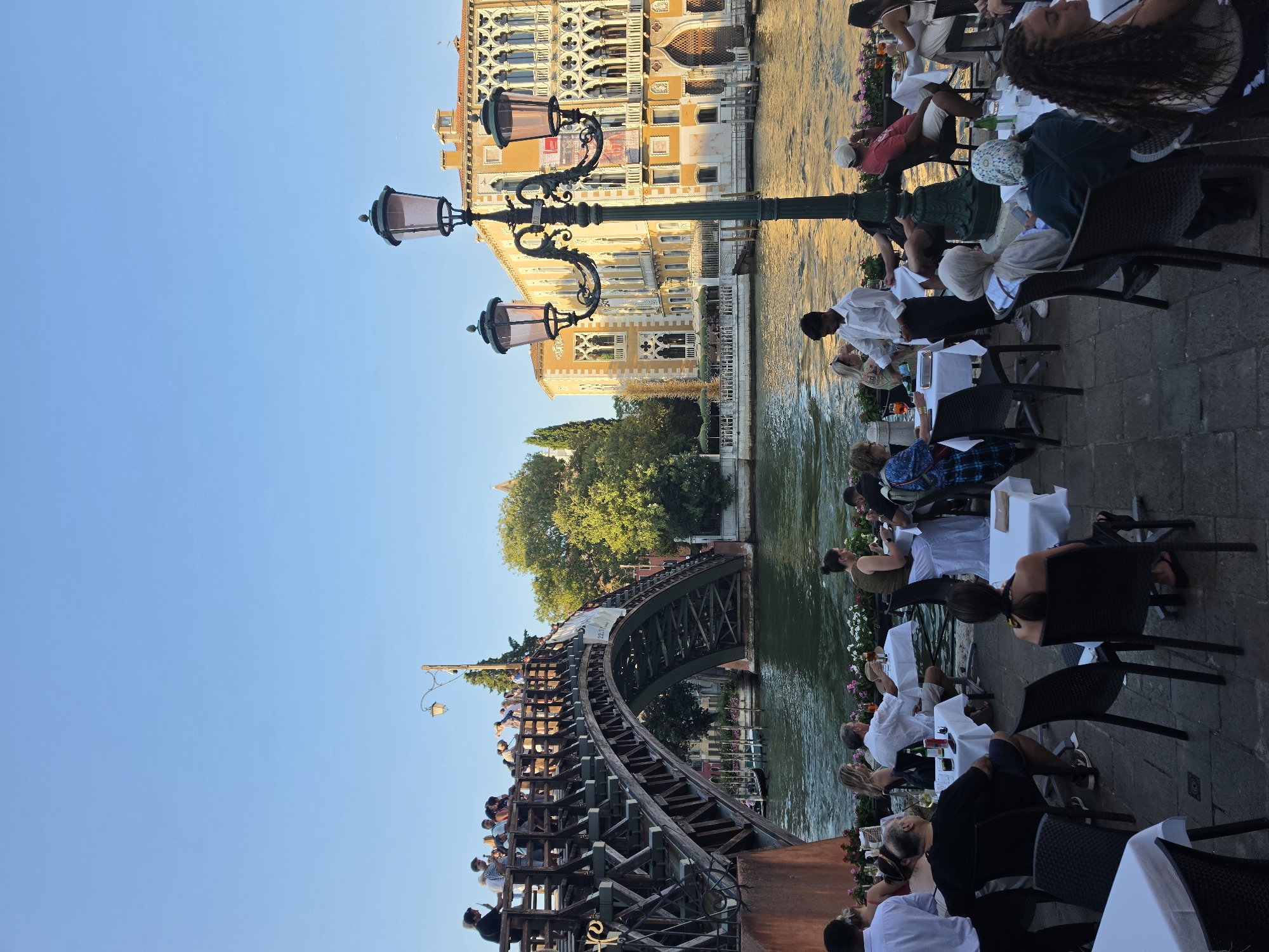 The image captures a lively outdoor dining scene in Venice, Italy, situated along a canal. In the foreground, several tables are set with white tablecloths and chairs, occupied by diners enjoying their meals and drinks. A waiter in a white shirt is seen serving a table.
Dominating the background is a picturesque wooden bridge, likely the Ponte dell'Accademia, with numerous people standing on it, observing the surroundings. Beyond the bridge and canal, traditional Venetian buildings with ornate architecture, painted in shades of yellow and orange, are visible. The canal itself reflects the warm hues of the buildings and the sky, creating a serene ambiance. Three ornate street lamps, characteristic of Venetian street furniture, are positioned in the scene, adding to the historical charm. The overall atmosphere is one of relaxed enjoyment amidst a beautiful and iconic Venetian cityscape.