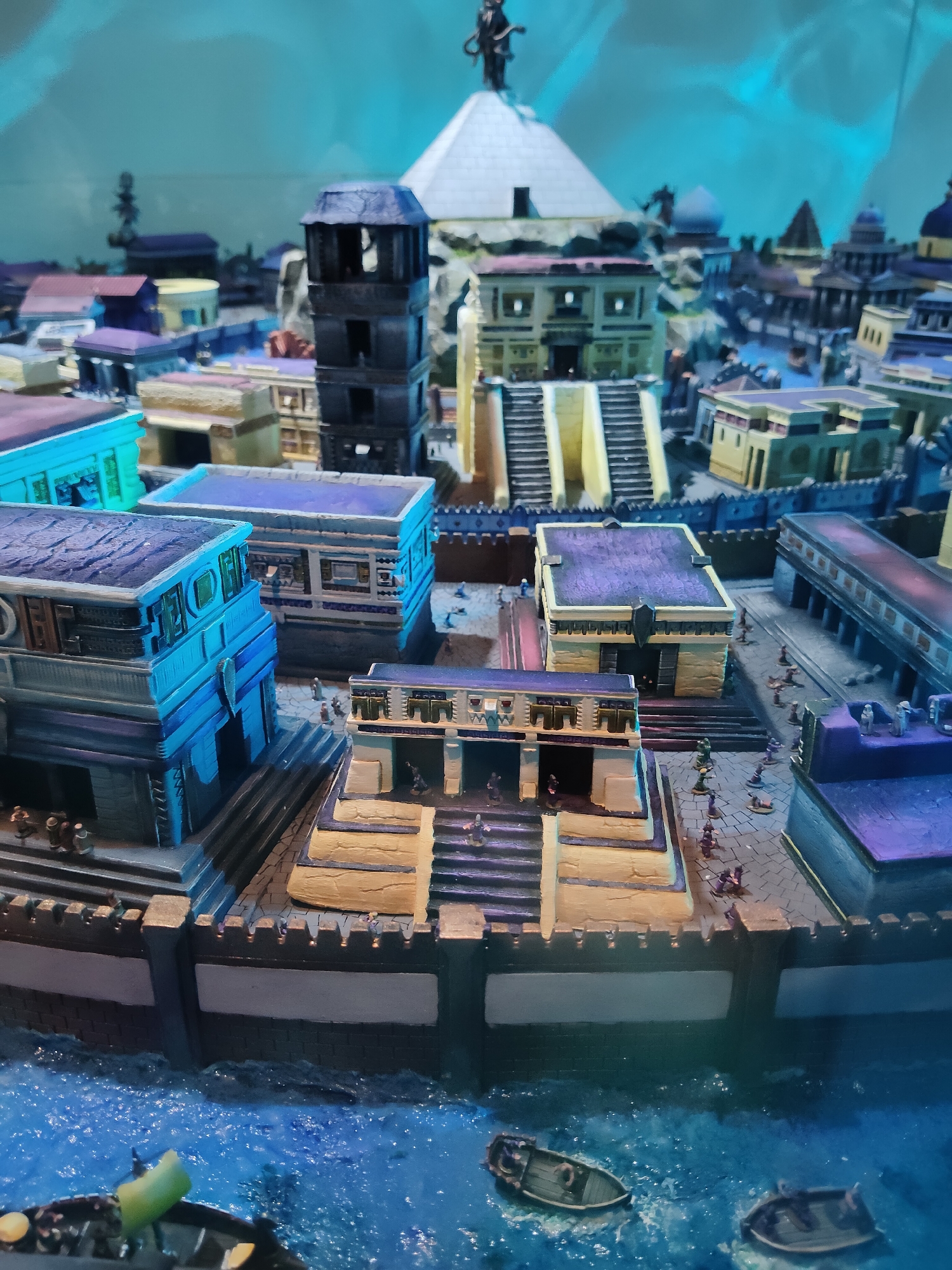 This is a close-up, eye-level, medium shot of a miniature model of a Mesoamerican city, likely representing Tenochtitlan or a similar ancient settlement. The model is positioned in a display with a blue, wavy backdrop, suggesting water or a stylized sky. The foreground features a section of blue, textured water with two small, dark-colored boats. One boat, closer to the viewer and on the left, has a visible green object protruding from its stern and a yellow circular object. The other boat, slightly further away and to the right, also appears to be occupied by small figures.
The city model itself is detailed and multi-leveled, with structures constructed from various colored blocks representing buildings, pyramids, and walls. In the center, a prominent light-colored stepped pyramid with a darker rectangular structure on top is a focal point. Surrounding it are several other buildings in shades of blue, purple, yellow, and tan, exhibiting architectural styles with stepped designs, staircases, and colonnades. Small, dark figures are scattered throughout the model, representing people engaged in various activities within the city. Some figures are on the steps of buildings, while others are walking along pathways or standing near structures. A dark, tall tower stands to the left of the central pyramid. The city is enclosed by a dark, fortress-like wall with crenellations, which separates the urban area from the water. The overall impression is that of a meticulously crafted diorama depicting an ancient civilization. The image does not provide any specific information or context linking it to the city of Singapore.