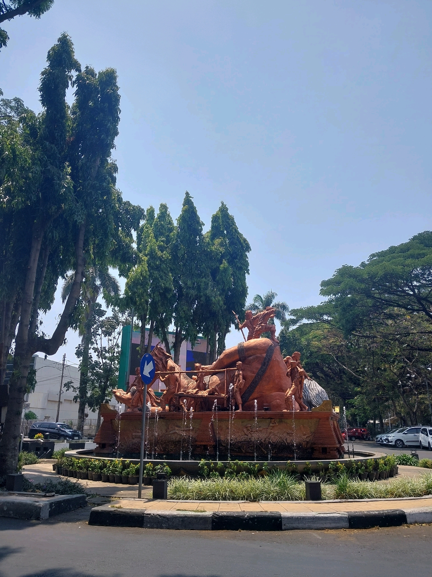 The image shows a wide shot of the Tugu Air Mancur fountain in Malang, Indonesia. The fountain is a large, ornate structure made of reddish-brown material, depicting several figures in dynamic poses, seemingly engaged in a struggle or a procession, pulled by a chariot-like vehicle. Water sprays from various points of the fountain, adding a sense of movement and life. The fountain is situated in a traffic circle, with roads and cars visible in the background. Tall, slender trees with lush green foliage line the sides of the road and surround the fountain area. The sky is a clear, pale blue, suggesting a sunny day. Several parked cars are visible on the right side of the image. A blue directional sign pointing left is mounted on a pole in front of the fountain. The overall scene depicts a well-maintained public space in an urban environment.