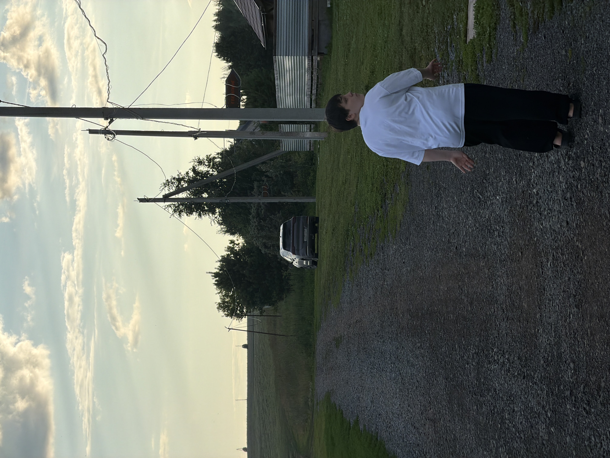 A young person with dark hair is standing outdoors, facing away from the camera. They are wearing a white long-sleeved shirt and black pants. Their arms are outstretched to their sides, with palms facing upwards. The person appears to be in a relaxed or contemplative pose.
The scene is a rural or semi-rural setting in Bol'shaya Ven'ya, Russia. In the background, there are utility poles with wires, a dark-colored SUV, and a building with a corrugated metal roof. Trees and a grassy field are also visible. The ground in the foreground is covered with gravel. The sky is light blue with scattered white clouds, suggesting it is daytime, possibly late afternoon or early evening. The overall atmosphere is calm and serene. There is no visible text in the image.
