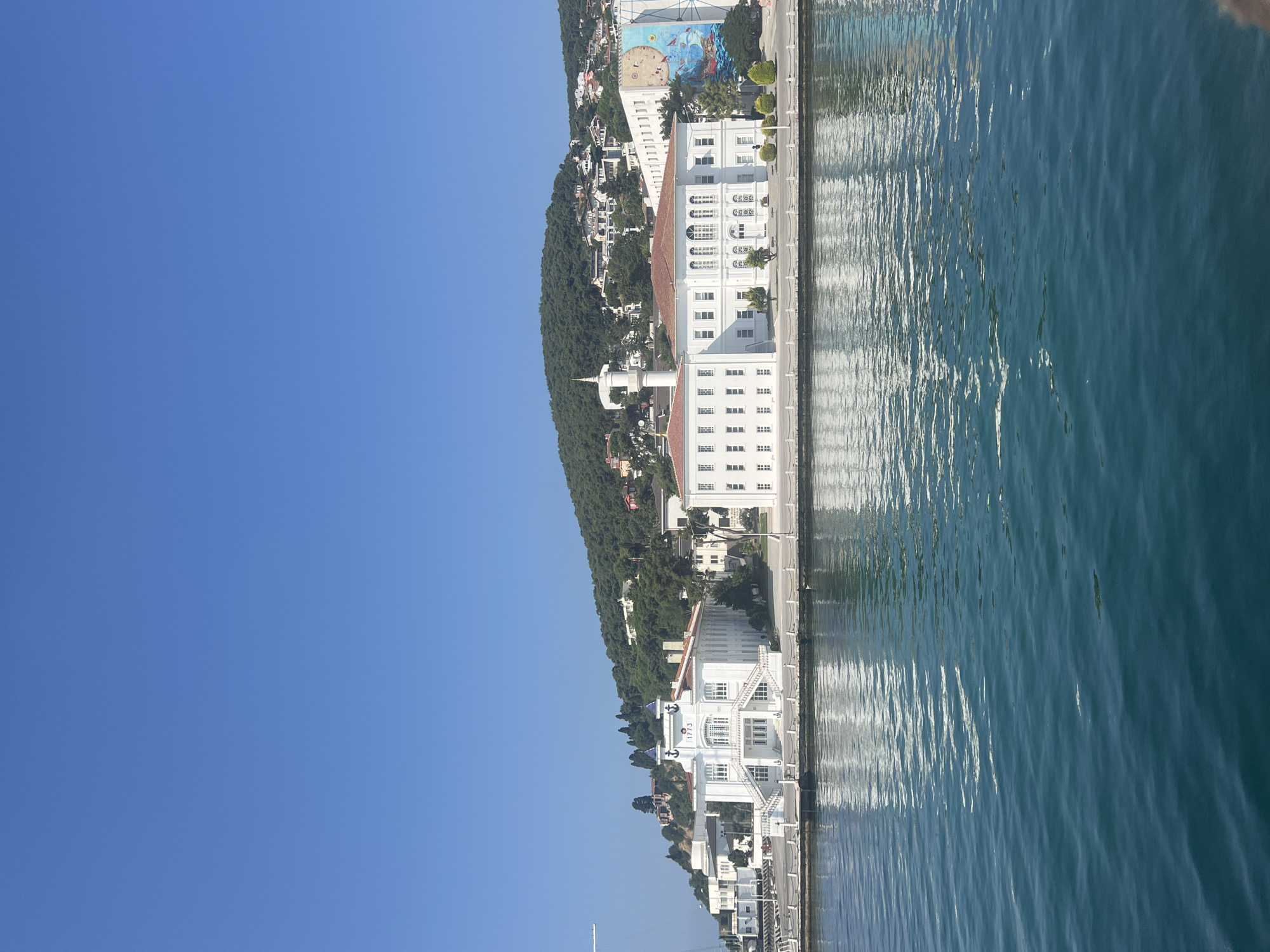 This is a landscape photograph taken from the water, looking towards a waterfront with several white buildings and a tree-covered hill in the background. The sky is clear and blue. The water in the foreground is a deep blue-green, with gentle waves reflecting the sky and the buildings.
The buildings are predominantly white with red-tiled roofs. The leftmost building has a symmetrical facade with a prominent entrance featuring wide, white stairs. It is adorned with two small blue cupolas topped with white spheres and an anchor emblem above the entrance. The year "1773" is visible above the anchor. This building appears to be the Khedive Palace, located on Heybeliada in the Princes' Islands, Turkey.
To the right of the Khedive Palace, a larger white building extends horizontally along the waterfront. This building has multiple windows and a central section with arched windows. To the far right of this building, a large, colorful mural is visible on the side of another white building. The mural depicts a blue moon and a nautical scene with ships and sea creatures.
Behind the buildings, a steep hill is covered in lush green trees and scattered houses with red roofs. The overall scene suggests a sunny day in a picturesque coastal area. No people are visible in the image. The composition emphasizes the architecture and the natural beauty of the location.