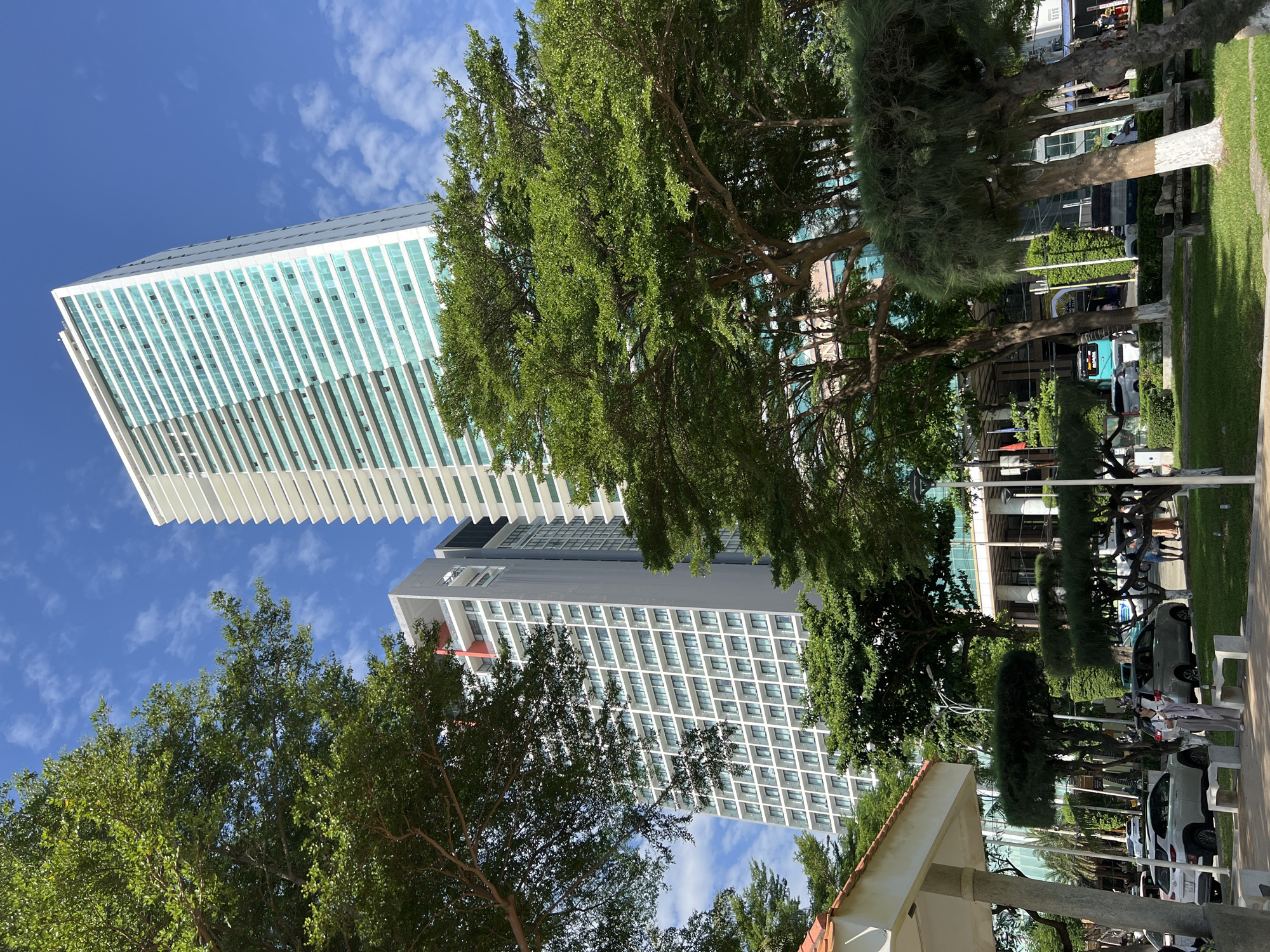 The image depicts a street scene in Nha Trang, Vietnam, characterized by modern high-rise buildings, lush trees, and parked cars under a bright blue sky with scattered clouds. Two prominent skyscrapers dominate the background. The building on the right is a sleek, white high-rise with numerous floors, featuring a facade of alternating white panels and balconies with teal-tinted glass windows. To its left, another, slightly shorter skyscraper stands, predominantly grey with a grid of windows and some balconies visible.
In the foreground, several mature trees with full green canopies are strategically placed, providing shade and a natural contrast to the urban architecture. Their branches extend across the frame, partially obscuring the buildings behind them. Below the trees, a paved walkway and a grassy area are visible. Several cars are parked along the roadside, including a white SUV and a silver minivan. People are present in the scene, some walking near the parked cars and others appearing to be in the vicinity of the buildings. One person, a woman wearing striped pants and a white top, is standing in the middle of the frame, looking at her phone. Another individual, a person wearing a white shirt and blue pants, is walking on the right side of the image. Benches are scattered on the grass, and lampposts are also present. The environment suggests a well-maintained urban space, possibly a promenade or a public area adjacent to commercial or residential buildings.