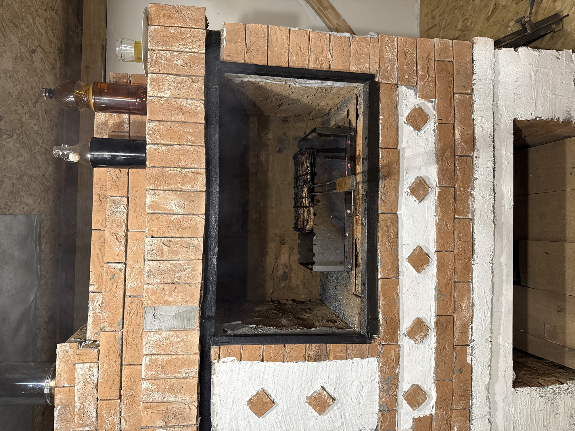 The image shows a brick barbecue oven located in Gon'ba, Russia. The oven has an open firebox containing a metal grill with food. On top of the oven, there are two bottles, one dark and one amber, and a glass with a yellow liquid. The oven is made of red bricks with white decorative elements and is situated indoors, with wooden flooring and ceiling visible. The overall scene suggests a setting for cooking and dining.