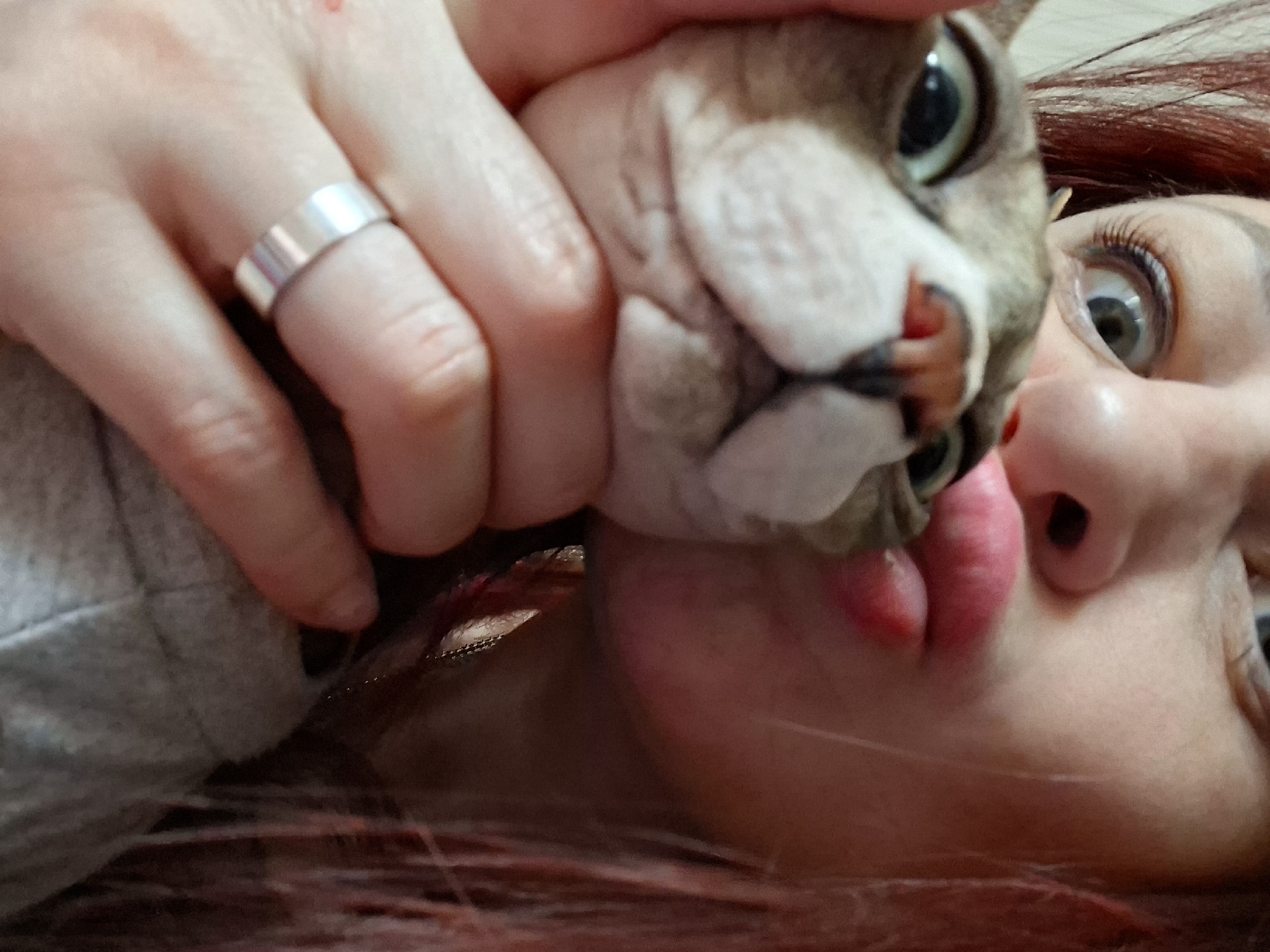 A close-up photograph depicts a person holding a Sphynx cat in close proximity to their face. The person's face, angled to the right, features red hair and a fair complexion. Their eyes are wide and appear to be looking upwards. The cat is pressed close to the person's mouth, with the cat's face prominently displayed. The person's hand, bearing a silver ring on one of the fingers, is partially visible. The background is out of focus and provides no clear indication of location. The image does not contain information to confirm it was taken in Забалоцкі сельскі Савет, Belarus.