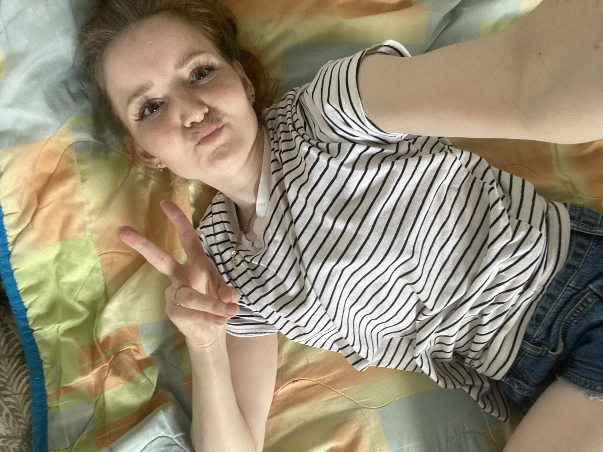 A young woman with light skin and light brown hair is shown in a selfie, lying on a colorful patterned bedspread in what appears to be an indoor setting. She is wearing a white and black striped short-sleeved top and blue denim shorts. Her head is positioned towards the upper left of the frame, and she is looking directly at the camera with her lips pursed in a slight pout. Her right hand is raised, making a peace sign (V-sign) with her index and middle fingers. She has visible eyelashes and a small earring in her right ear. Her left arm is extended upwards, suggesting she is holding the camera to take the selfie. The bedspread features a mix of light blue, orange, green, and yellow patches. The lighting suggests it might be daytime. There is no visible text in the image. The general atmosphere is relaxed and informal.