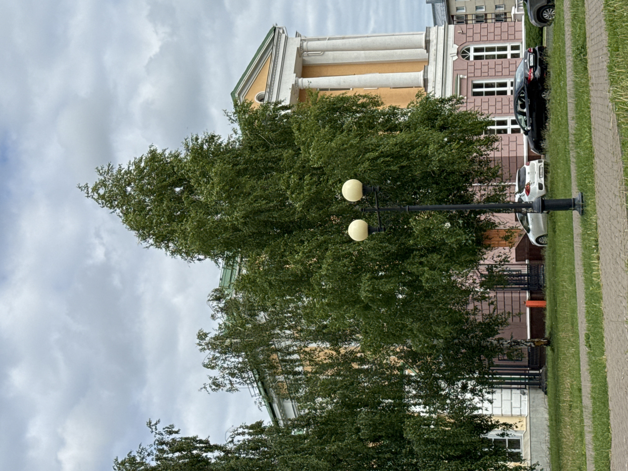 The image shows the exterior of a building with classical architectural elements, characterized by large columns, a pediment, and a green roof. The building facade is primarily painted in a pale orange and pink hue, with white trim around the windows and architectural features. Several large, leafy trees partially obscure the view of the building. In the foreground, there is a lamppost with two globe-shaped lights. Beyond the trees and building, there are parked cars on what appears to be a street or parking area. The sky is overcast with gray clouds.
This building is the Udmurt State Academic Theater in Izhevsk, Russia, located on Sovetskaya Street. The theater is a prominent cultural institution in the city.