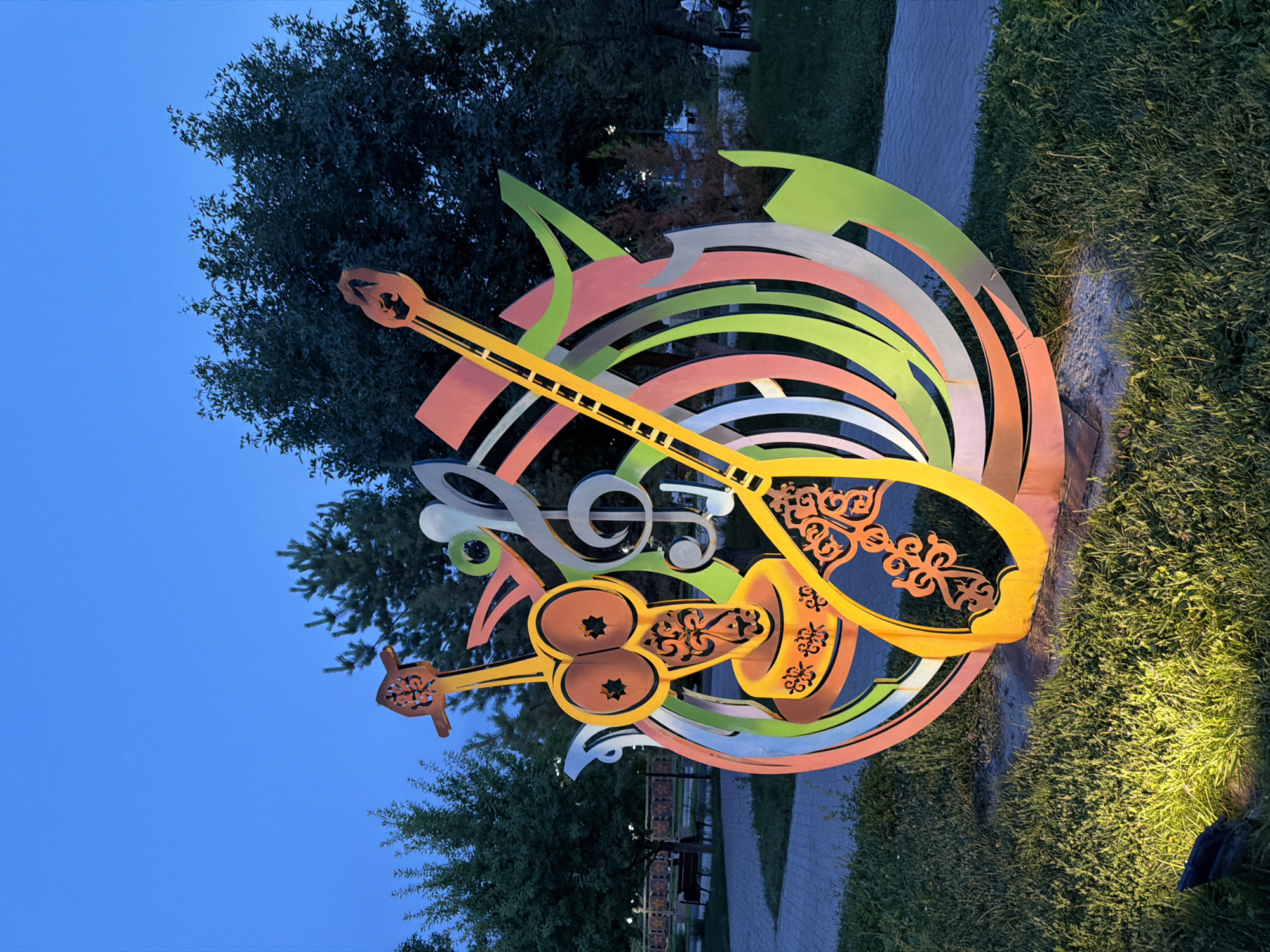 The image captures a vibrant, abstract sculpture in the city of Shymkent, Kazakhstan, likely at dusk or in the early evening. The sculpture, positioned on a grassy area next to a paved pathway, is composed of brightly colored metal elements arranged in swirling, circular patterns. Dominating the foreground is a stylized representation of musical instruments, including what appears to be a lute or a dombra, rendered in yellow with intricate orange decorative patterns. A treble clef symbol is also incorporated into the design. The sculpture is illuminated from below by a spotlight, casting a warm glow on the surrounding grass. In the background, trees form a dark backdrop against the twilight sky, and glimpses of park benches and distant figures suggest a public space. The overall impression is one of a dynamic and artistic installation within an urban park setting.