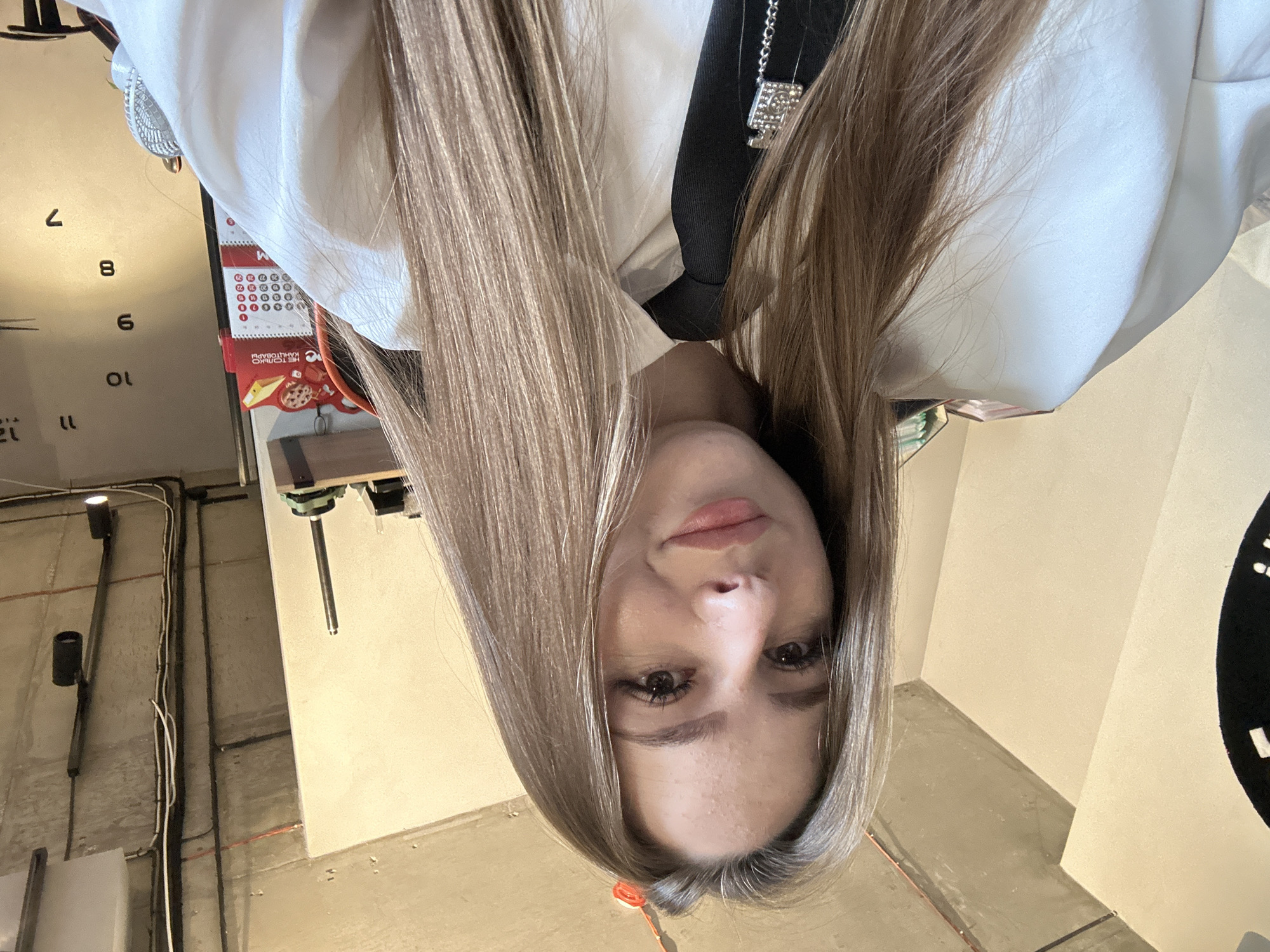 A young woman with long, straight, light brown hair, wearing a white collared shirt and a black tie with a decorative silver charm, is photographed from the chest up. Her eyes are looking towards the camera, and she is smiling slightly. The background appears to be an indoor setting with industrial-style concrete ceilings and exposed pipes. A large clock face is partially visible on the left, and a calendar is visible on the right.