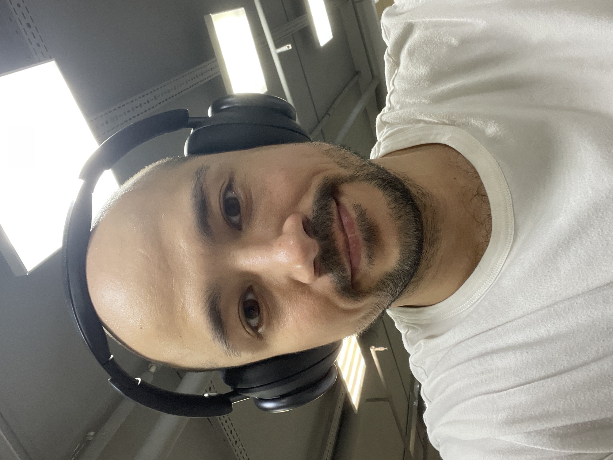 A man with a bald head and dark stubble wears black headphones and a white t-shirt. He is looking directly at the camera with a slight smile. Above him are industrial-style fluorescent lights, mounted on a gray, metal grid. The location is not identifiable as Izhevsk, Russia, from the visual information provided.